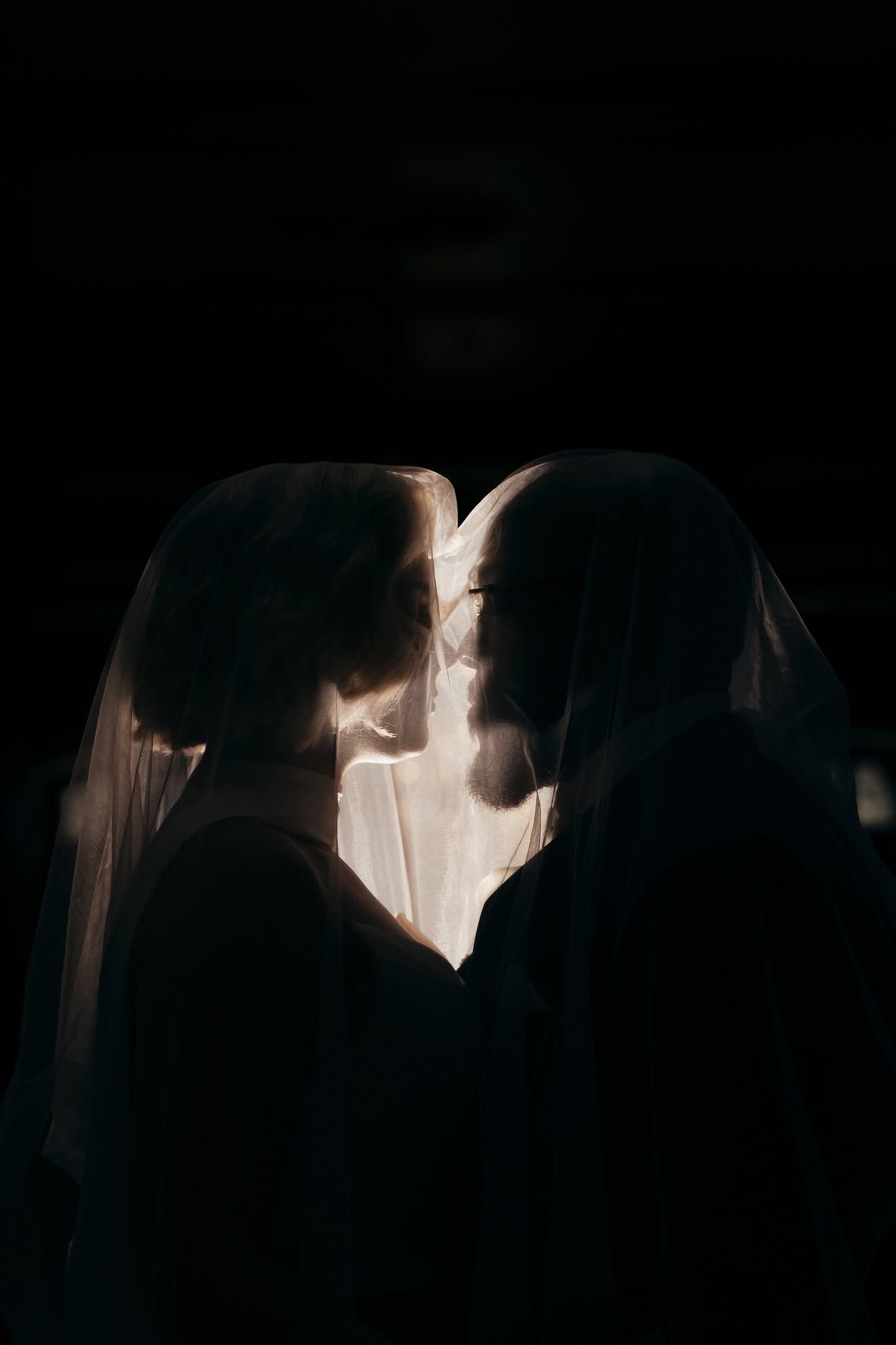 Romantic silhouette of a couple kissing under a bridal veil in dramatic dark lighting.