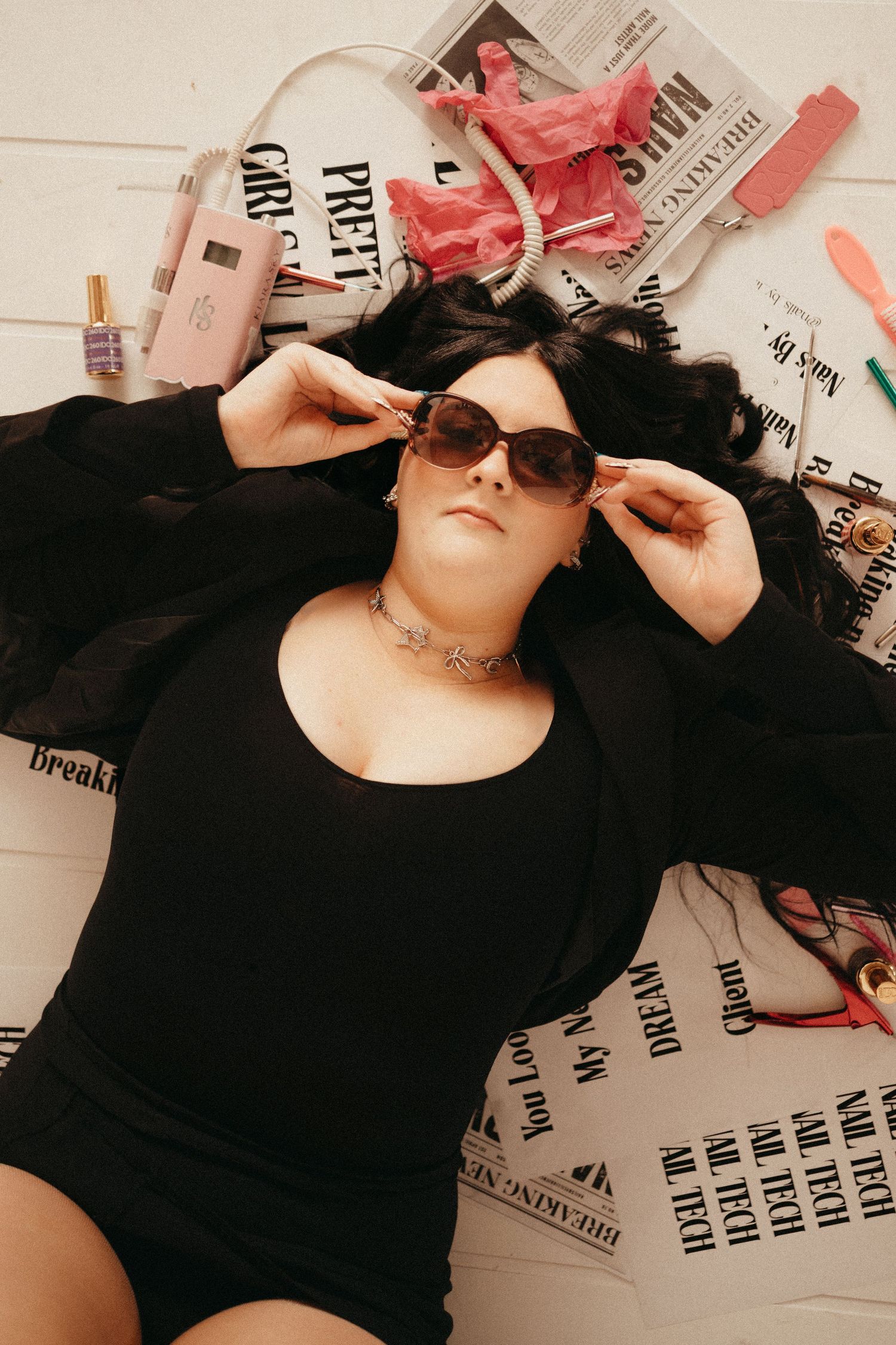 Woman in black bodysuit lying on newspaper print backdrop with beauty accessories.