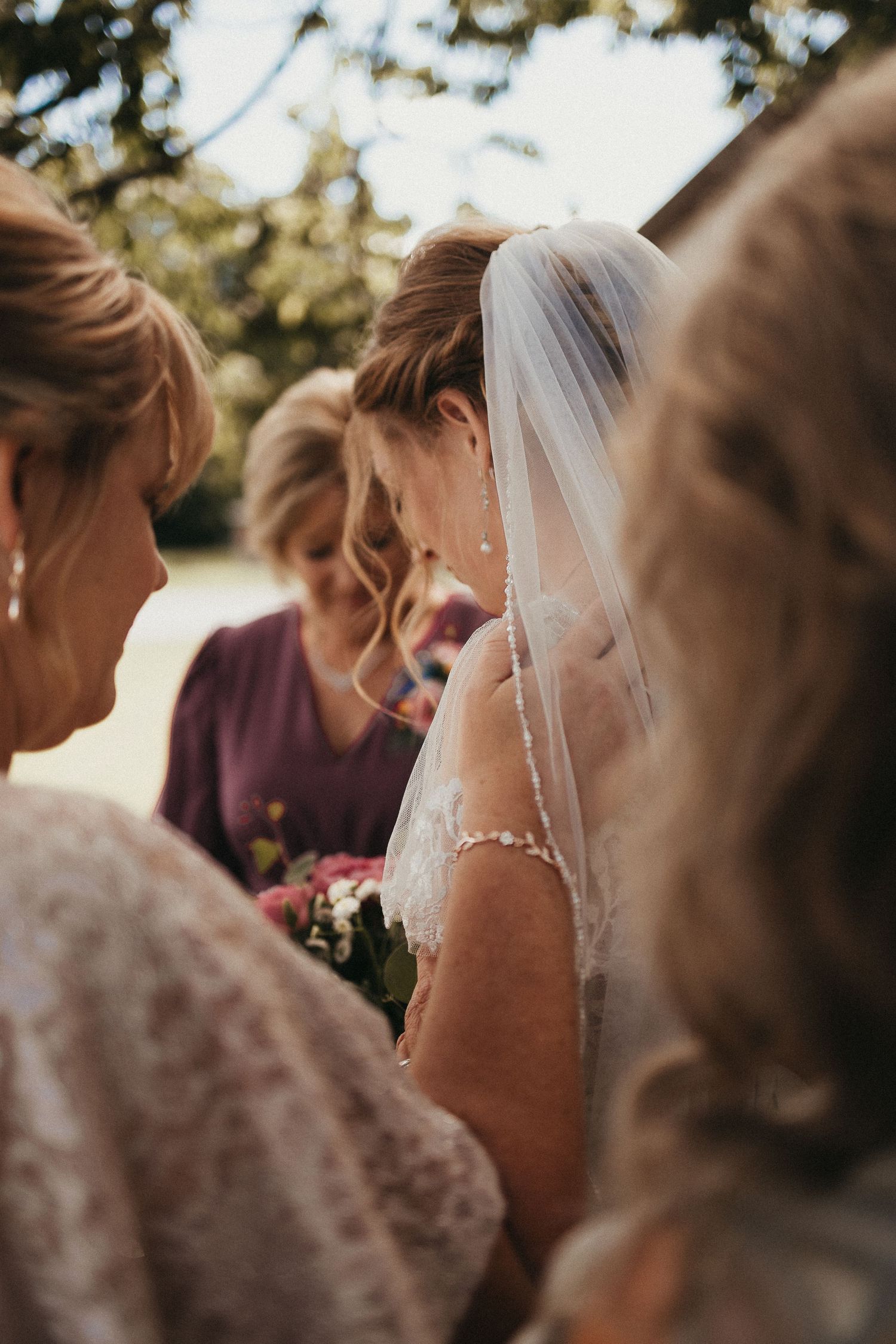 Bride in lace wedding gown and veil surrounded by bridesmaids in purple dresses outdoors.