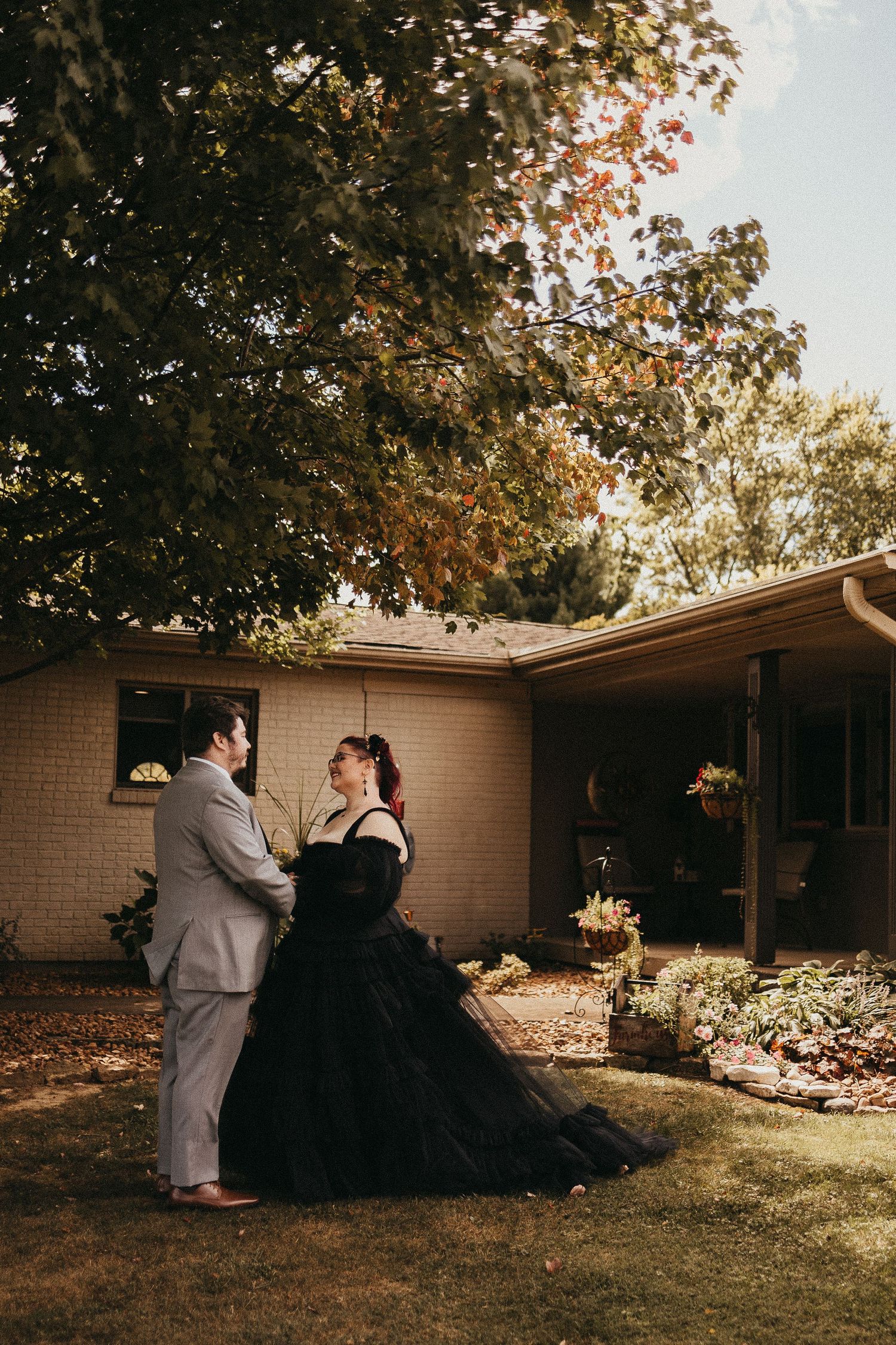 Couple shares a romantic moment in a sunlit backyard, man in gray suit and woman in a flowing black ball gown.