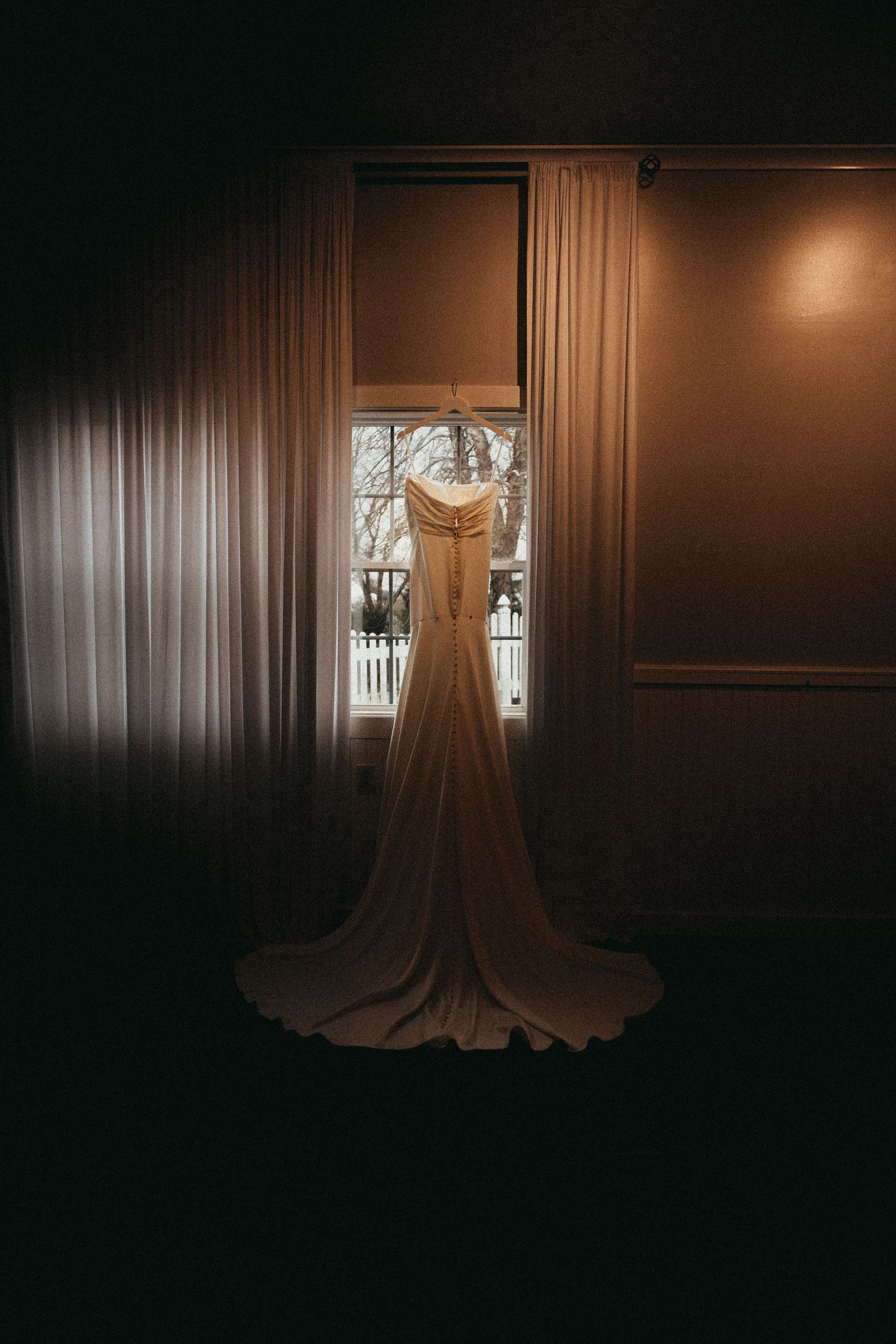 Elegant strapless white bridal gown with long train hanging in window before wedding ceremony.