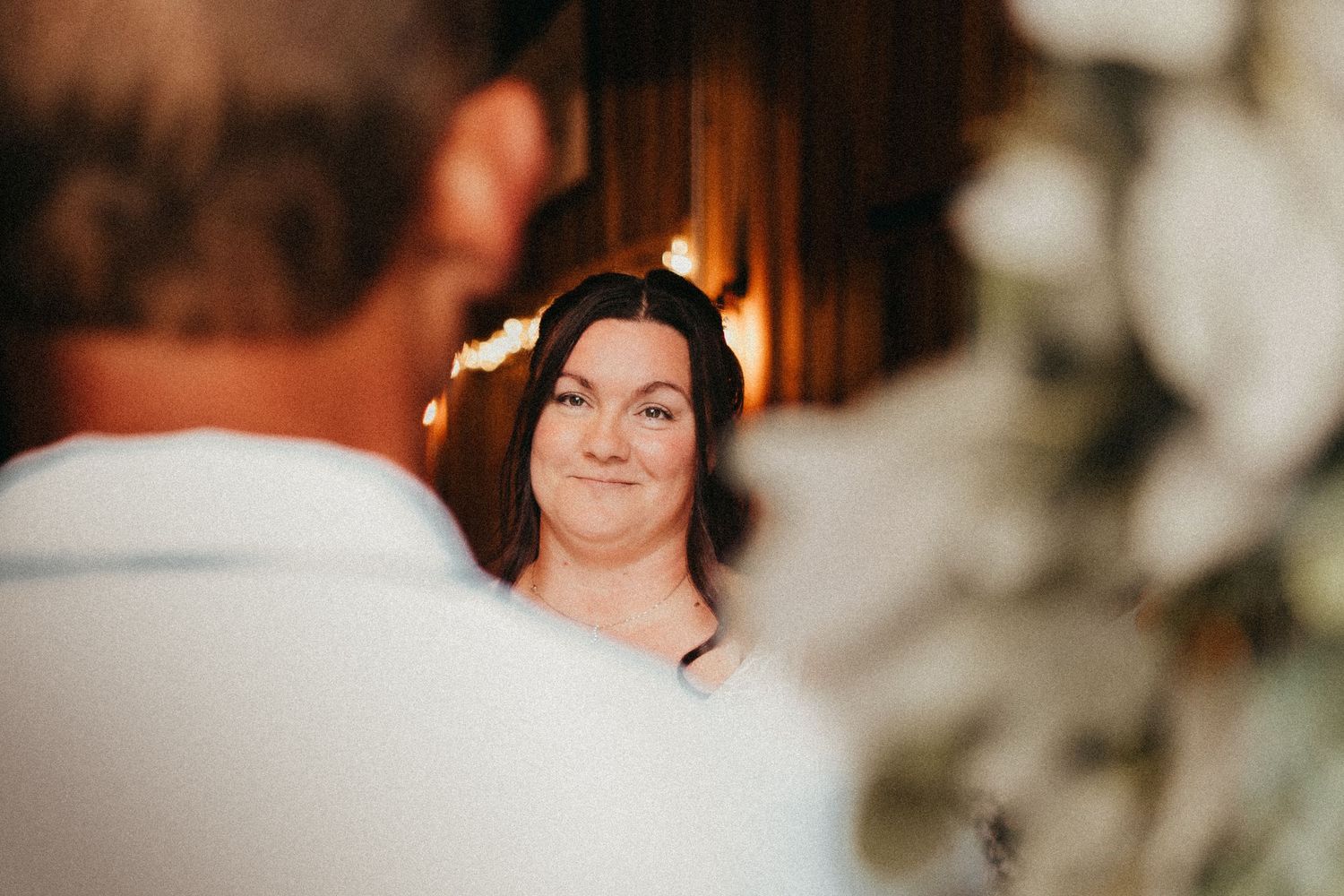 Emotional bride smiles lovingly at groom during intimate indoor wedding ceremony with warm lighting.