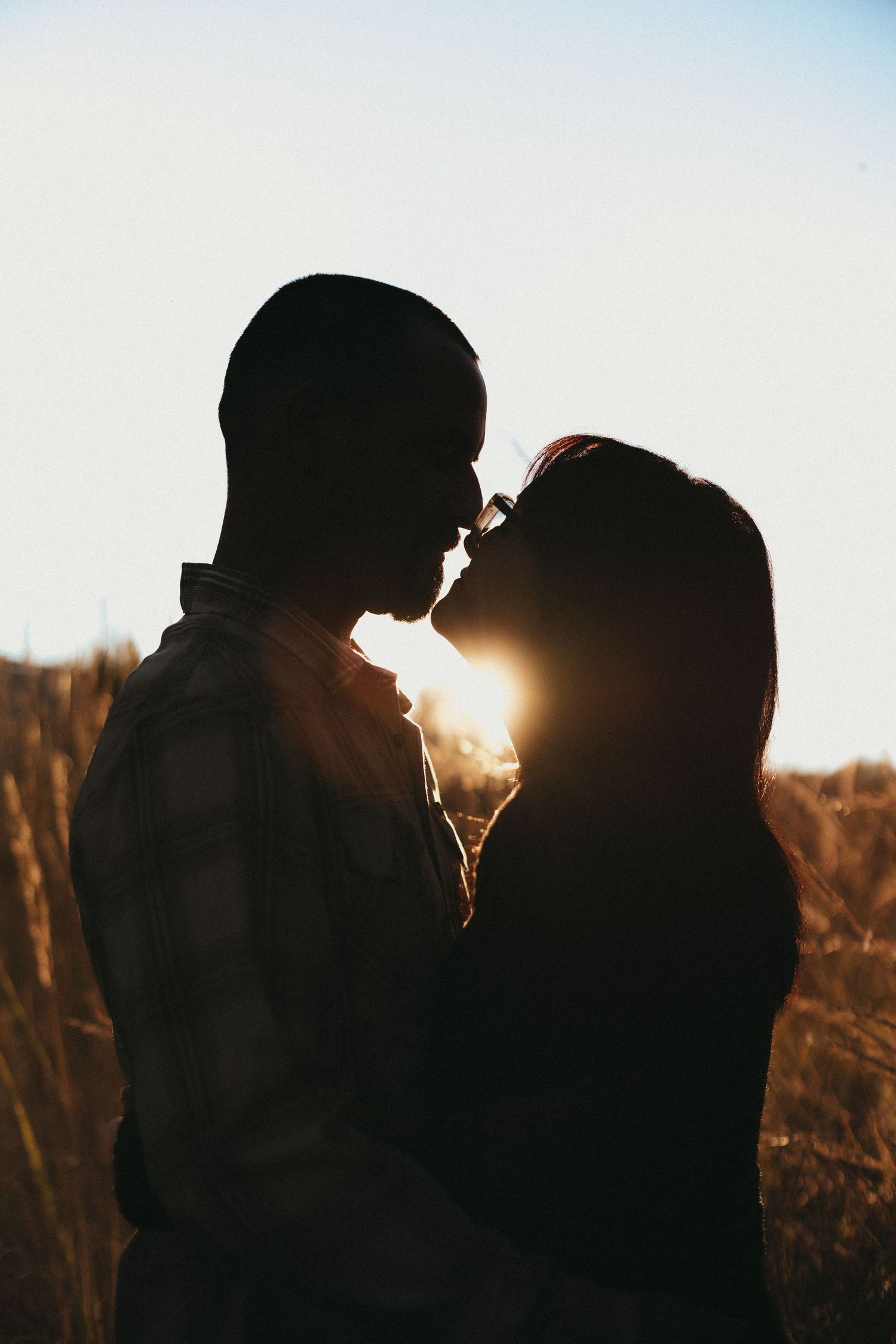 Romantic couple silhouette kissing at sunset in a golden field with warm backlight glowing between them.
