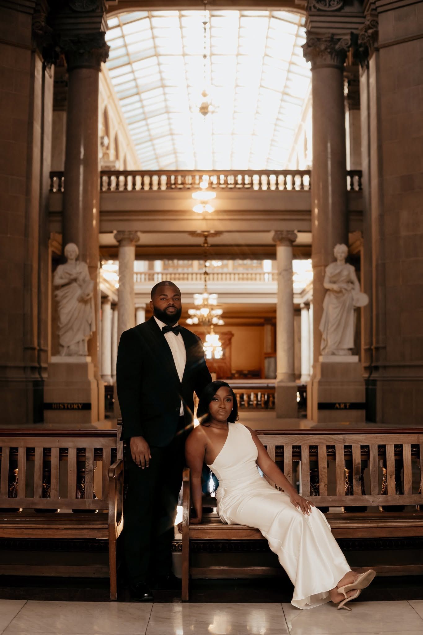 Elegant couple in formal attire posing in a grand hall with marble columns, statues, and a glass ceiling.