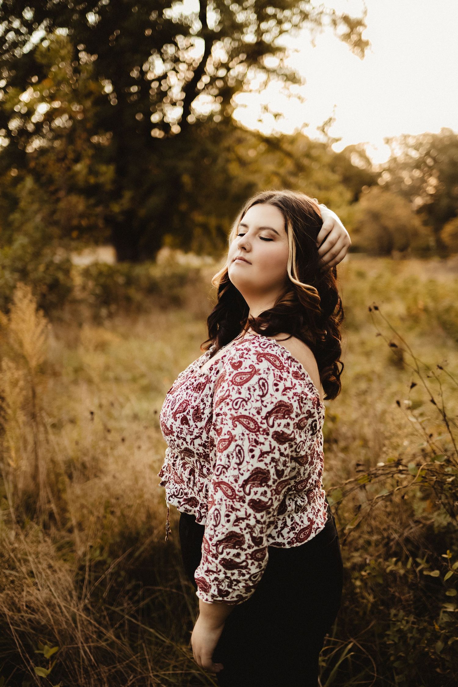 Young woman in floral top posing outdoors in a golden autumn field at sunset.