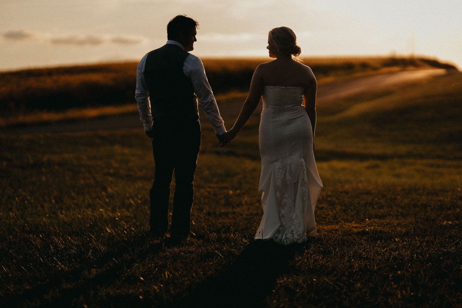 Bride and groom holding hands walking through a green field at golden hour sunset.