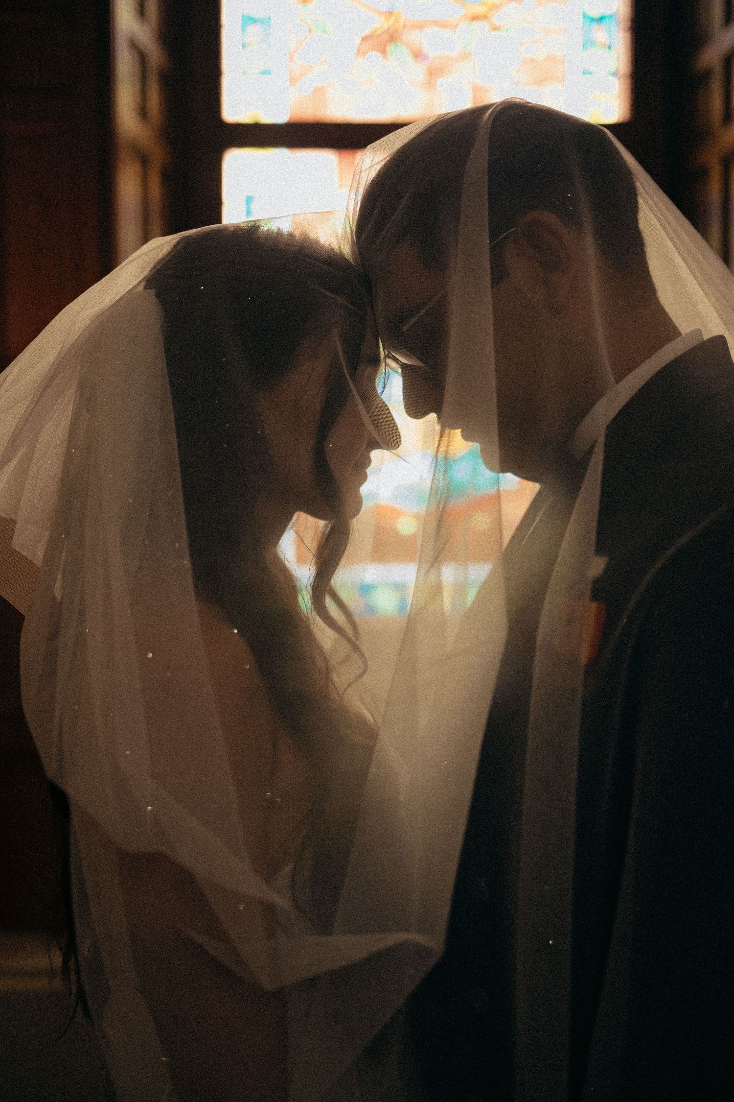 Bride and groom share an intimate moment under a veil, backlit by stained glass windows in a church.