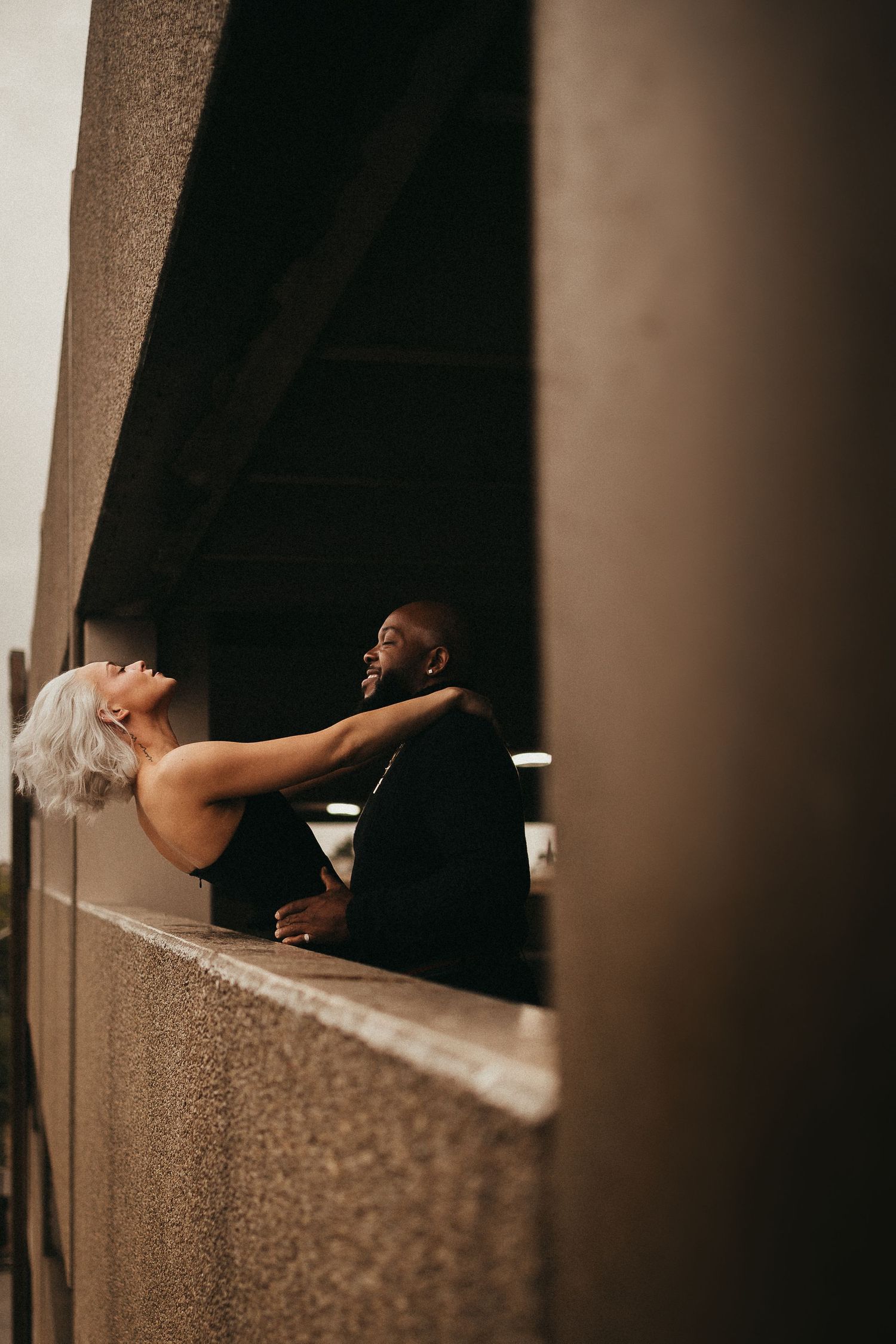 Interracial couple in a romantic embrace under a concrete overpass in moody urban setting.
