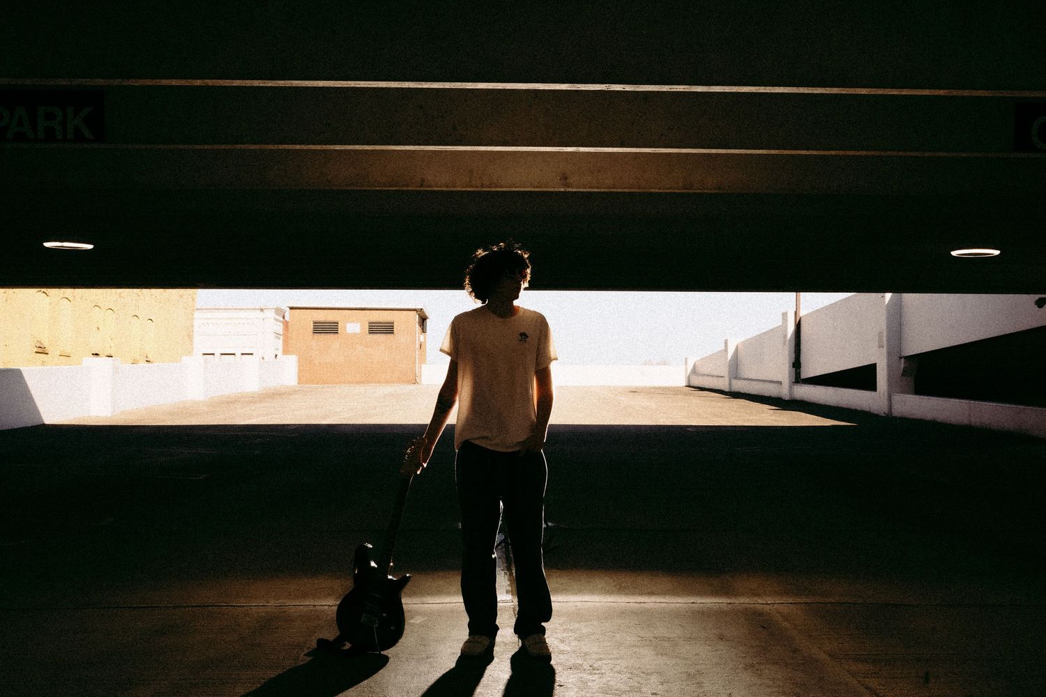Silhouette of person with guitar standing in shadowy parking garage entrance.