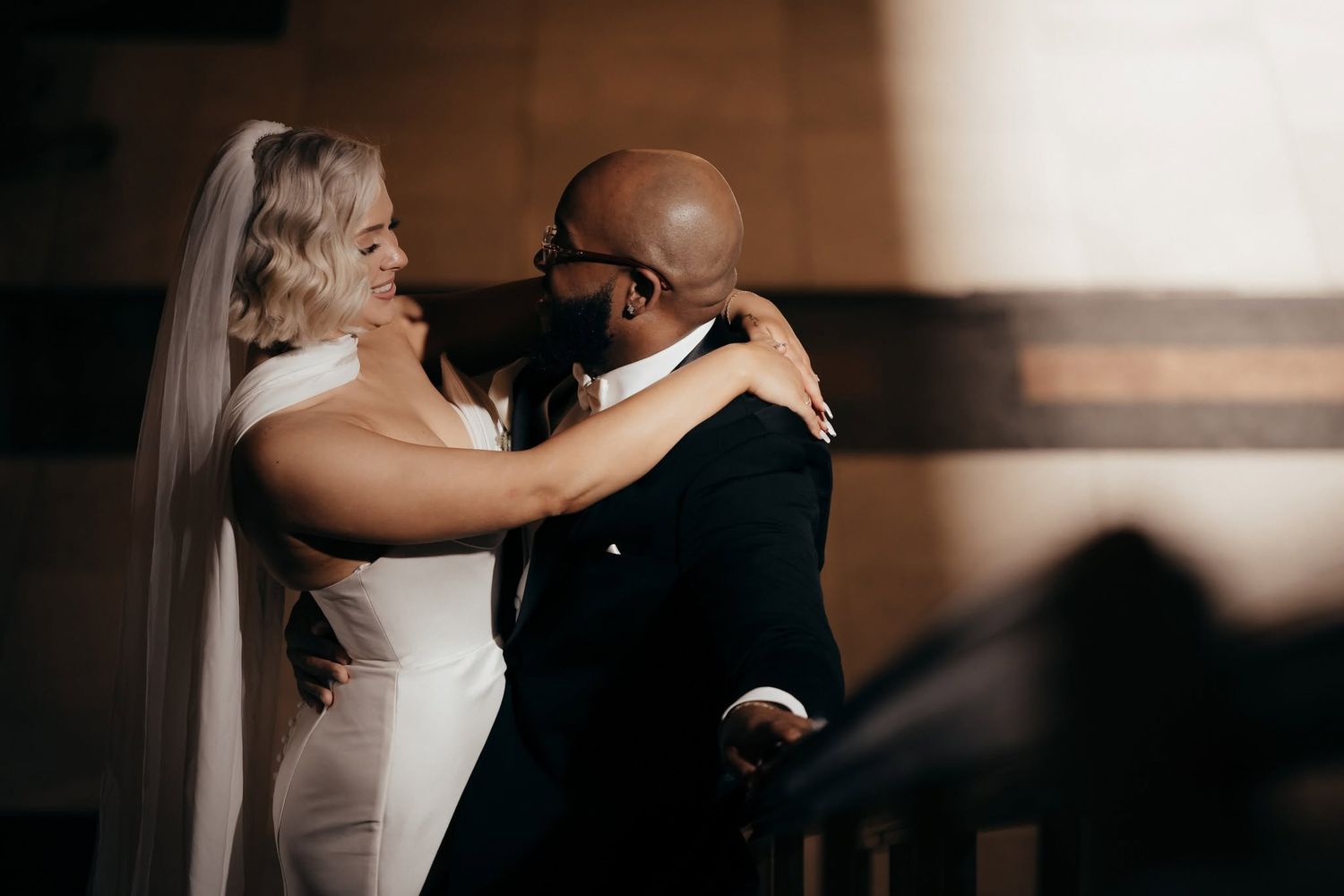 Bride and groom share an emotional first look embrace, wearing wedding dress with veil and dark suit.