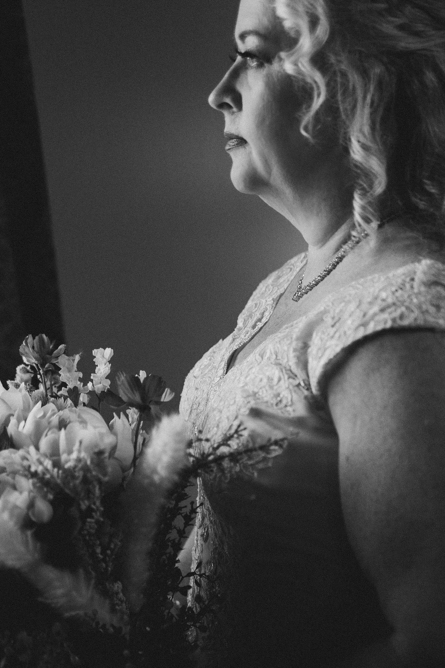 Black and white portrait of a bride in lace dress holding a bouquet, gazing thoughtfully to the side.