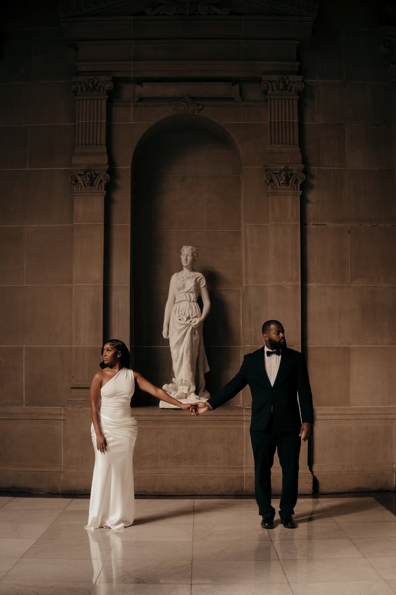 Elegant couple in wedding attire holding hands before a classical marble statue in a grand stone hall.