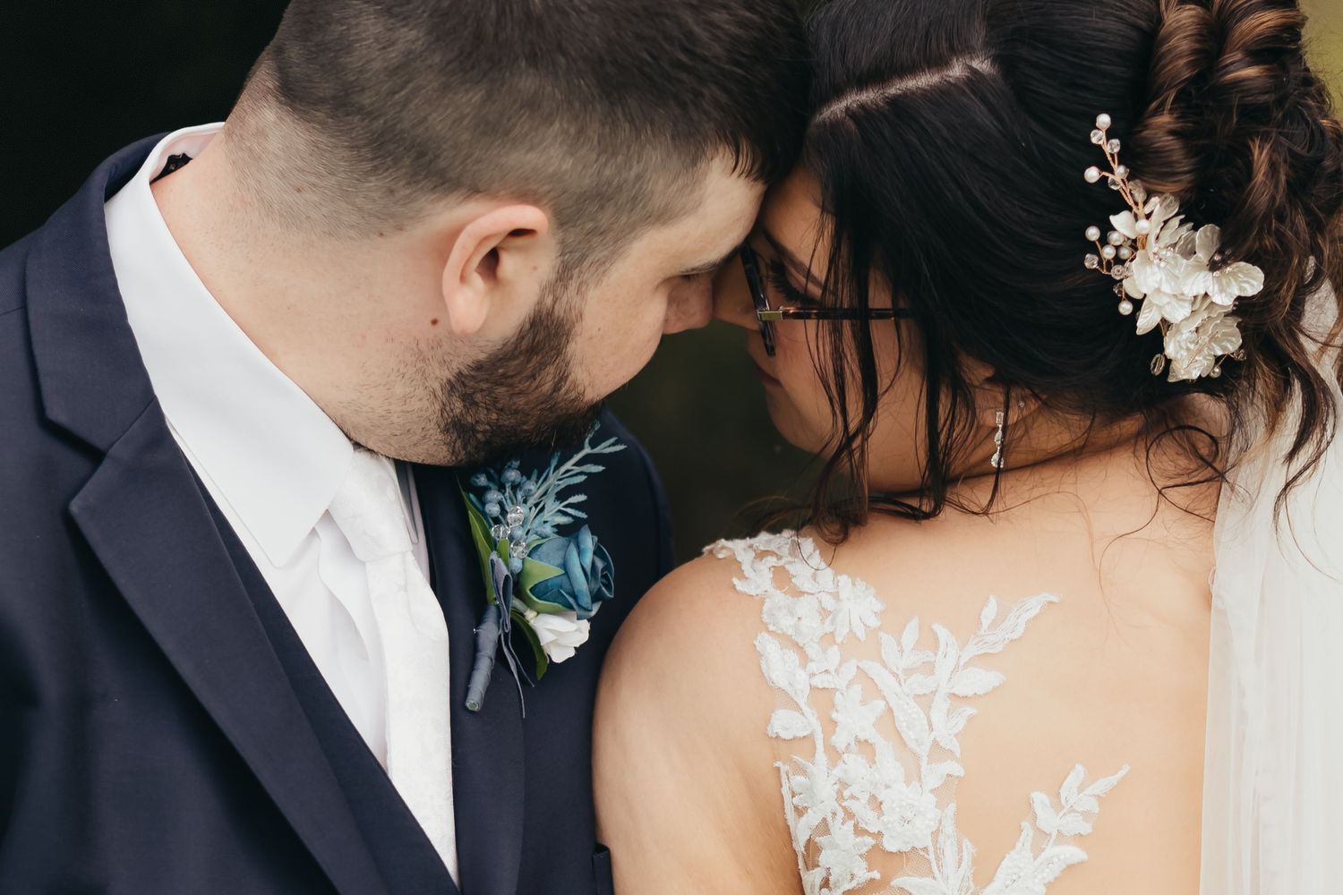 Bride and groom touching foreheads in romantic wedding portrait, bride wearing lace gown with floral hair accessory.