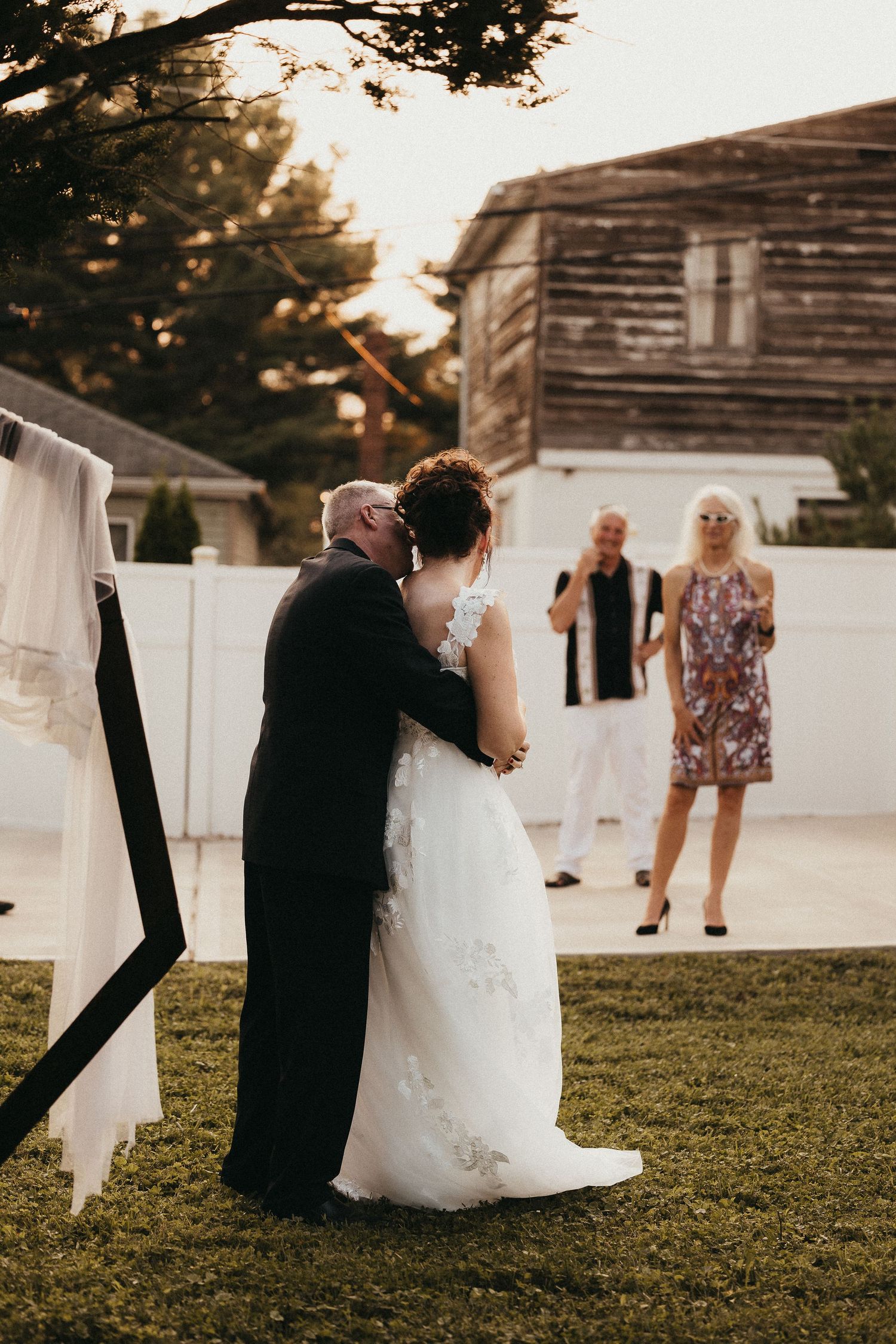 Bride and groom share first kiss during outdoor backyard wedding ceremony at golden hour with guests watching.