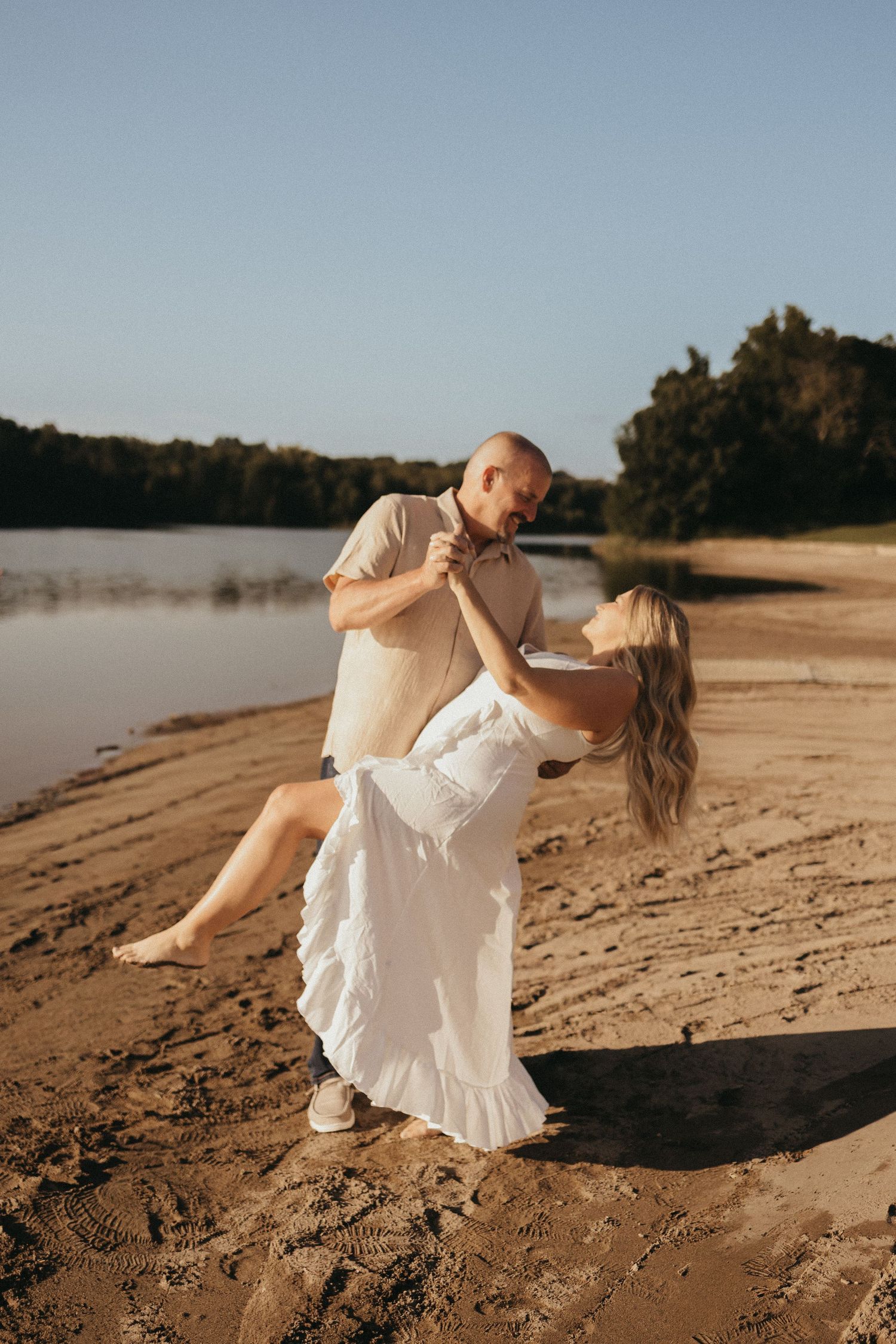 A couple dancing romantically on a sandy beach at golden hour, woman in white dress dipping gracefully by a calm river.