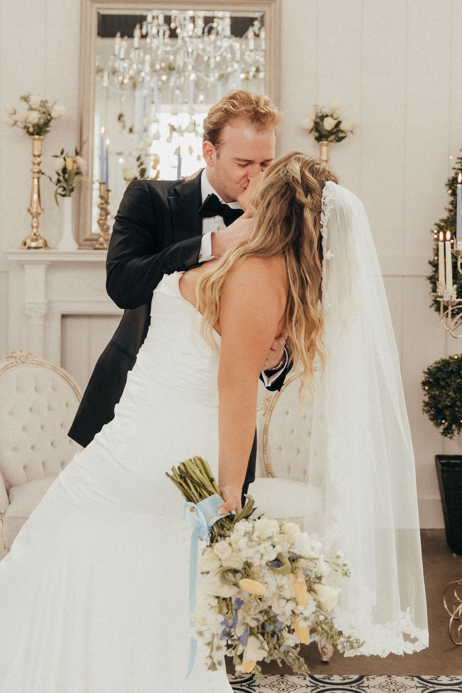Bride and groom share first kiss in elegant white venue with chandelier and floral decor.