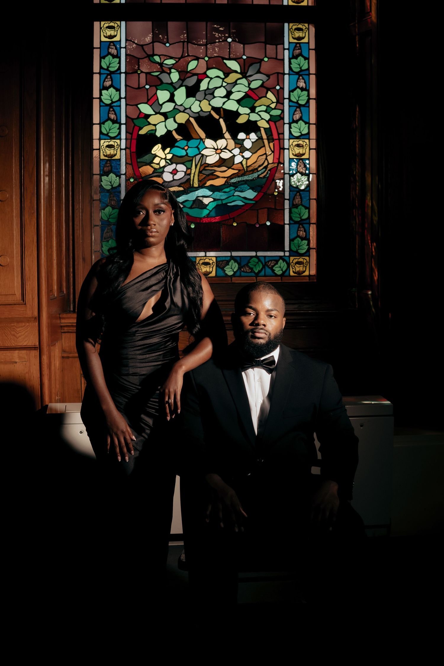 Elegant couple in formal black attire pose before a colorful stained glass window, the woman standing and the man seated in a bow tie.