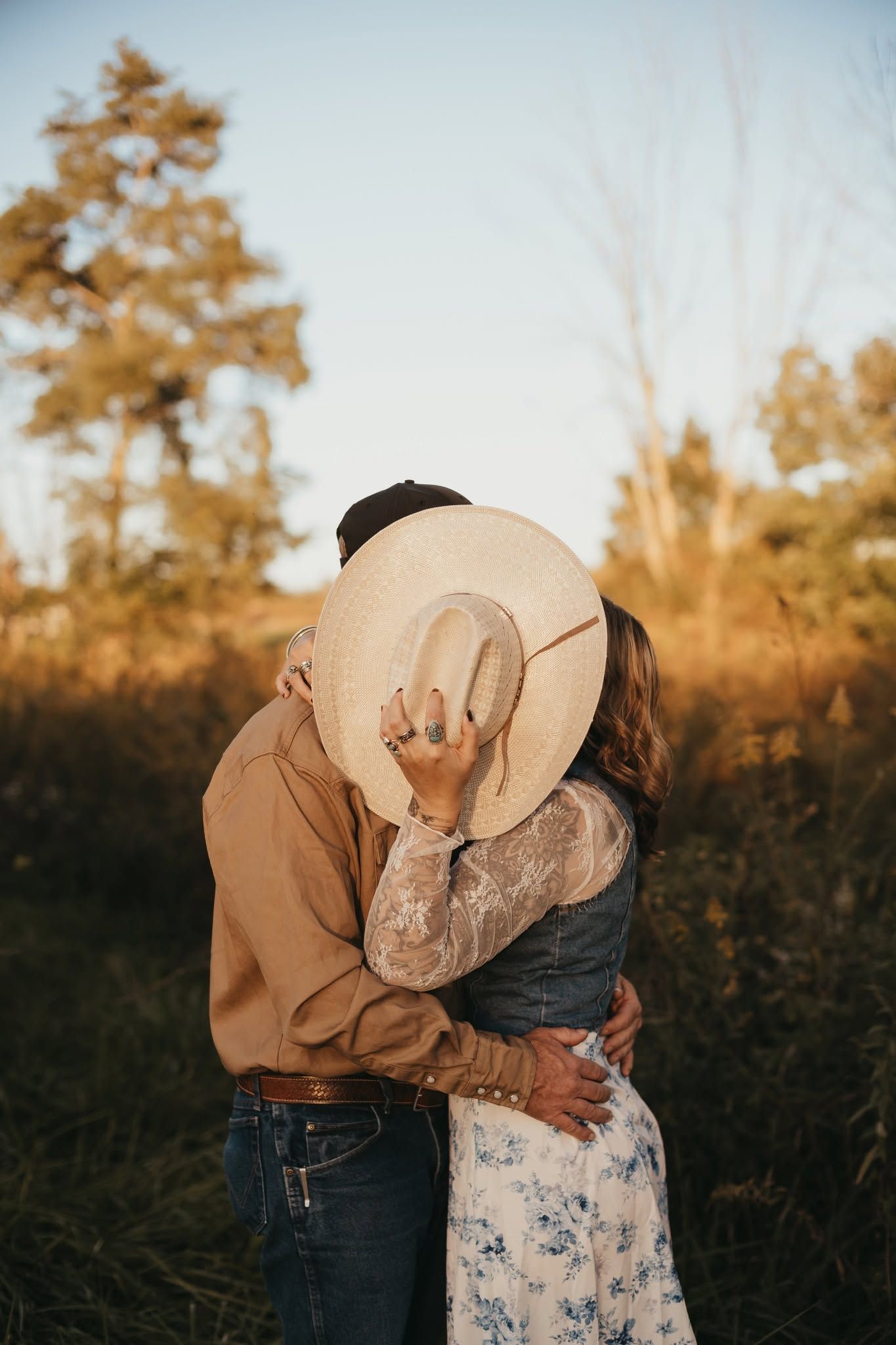 Couple embracing in golden hour light, faces hidden behind a wide-brim straw hat, surrounded by nature.