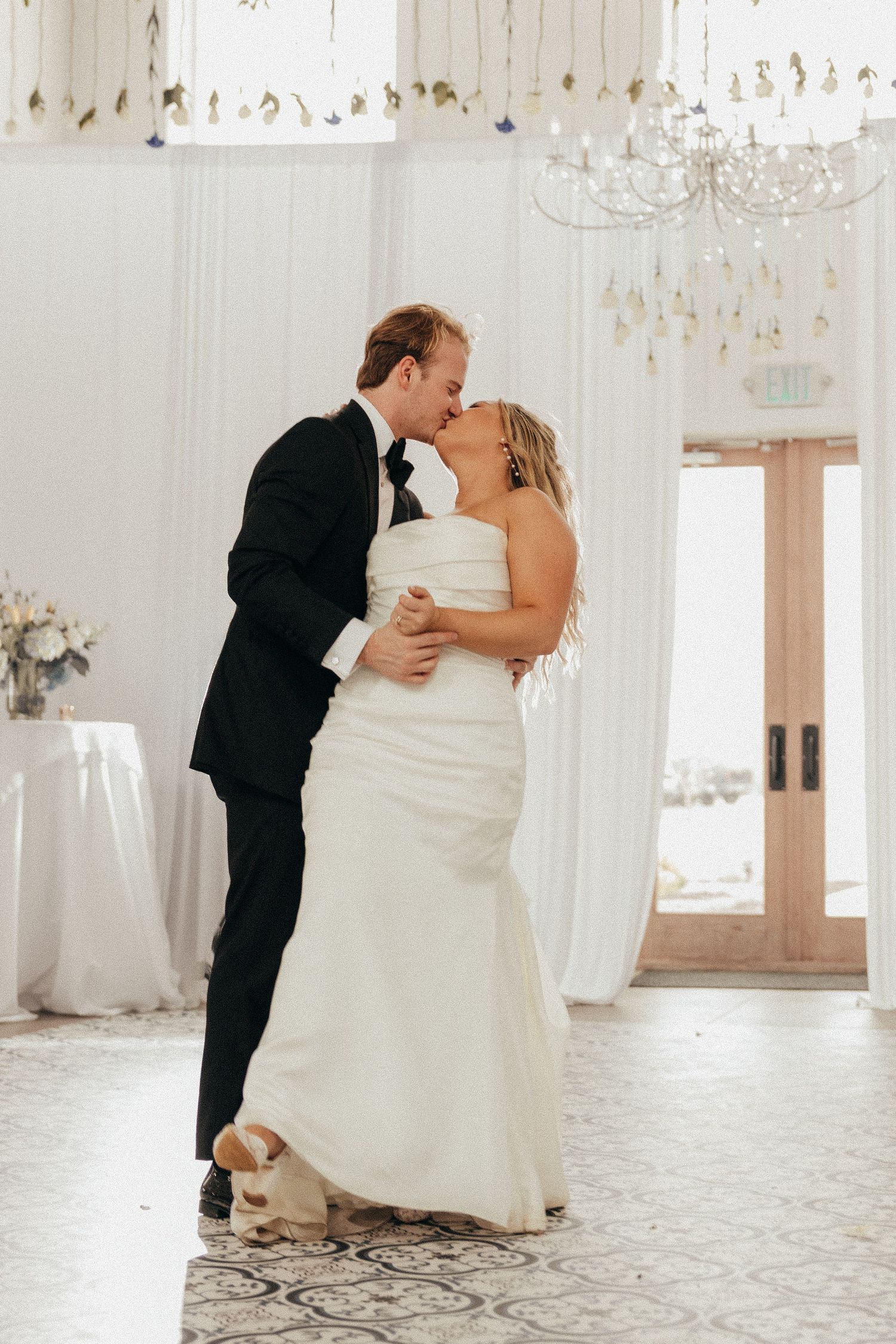 Bride and groom share their first dance kiss in an elegant white wedding reception venue with chandelier.