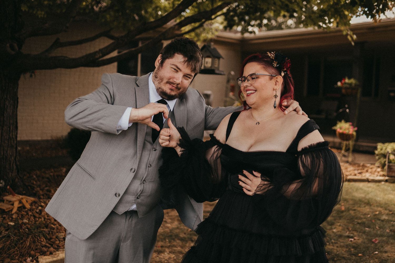 Couple laughs and poses playfully outdoors, man in gray suit and woman in a stunning black tulle off-shoulder gown.