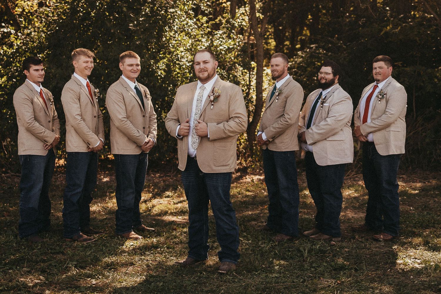 Groom and six groomsmen in matching tan blazers and dark jeans posing outdoors in sunlit wooded setting.