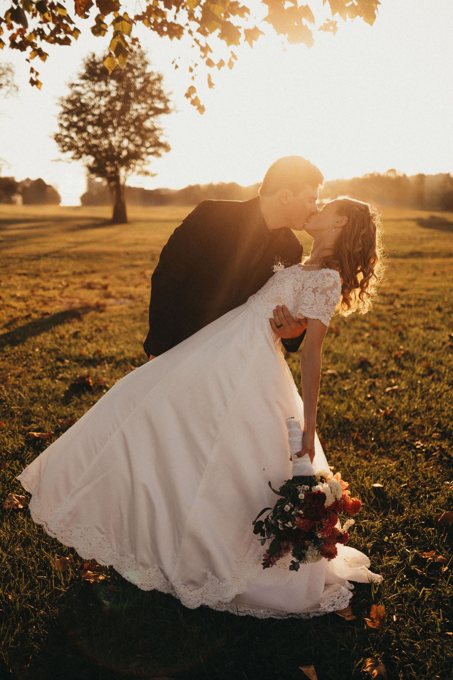 Groom dipping bride in white lace gown for a kiss during golden sunset in an open field.