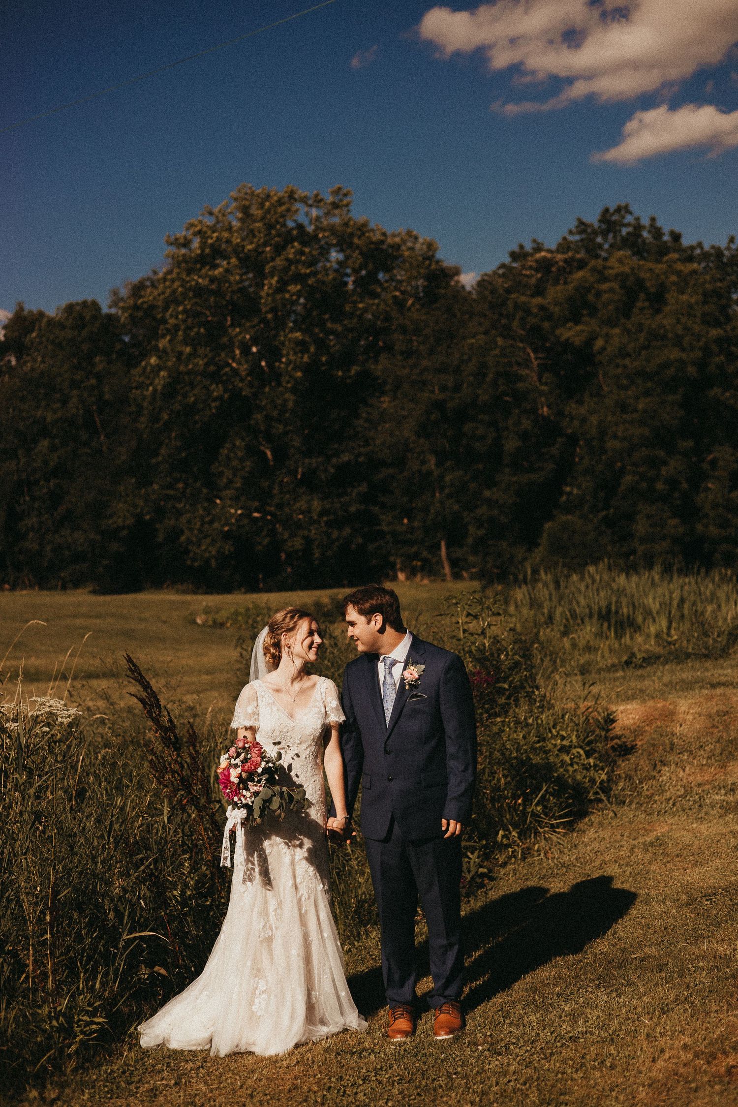 Bride and groom holding hands in a sunlit field with lush green trees and blue sky on their wedding day.