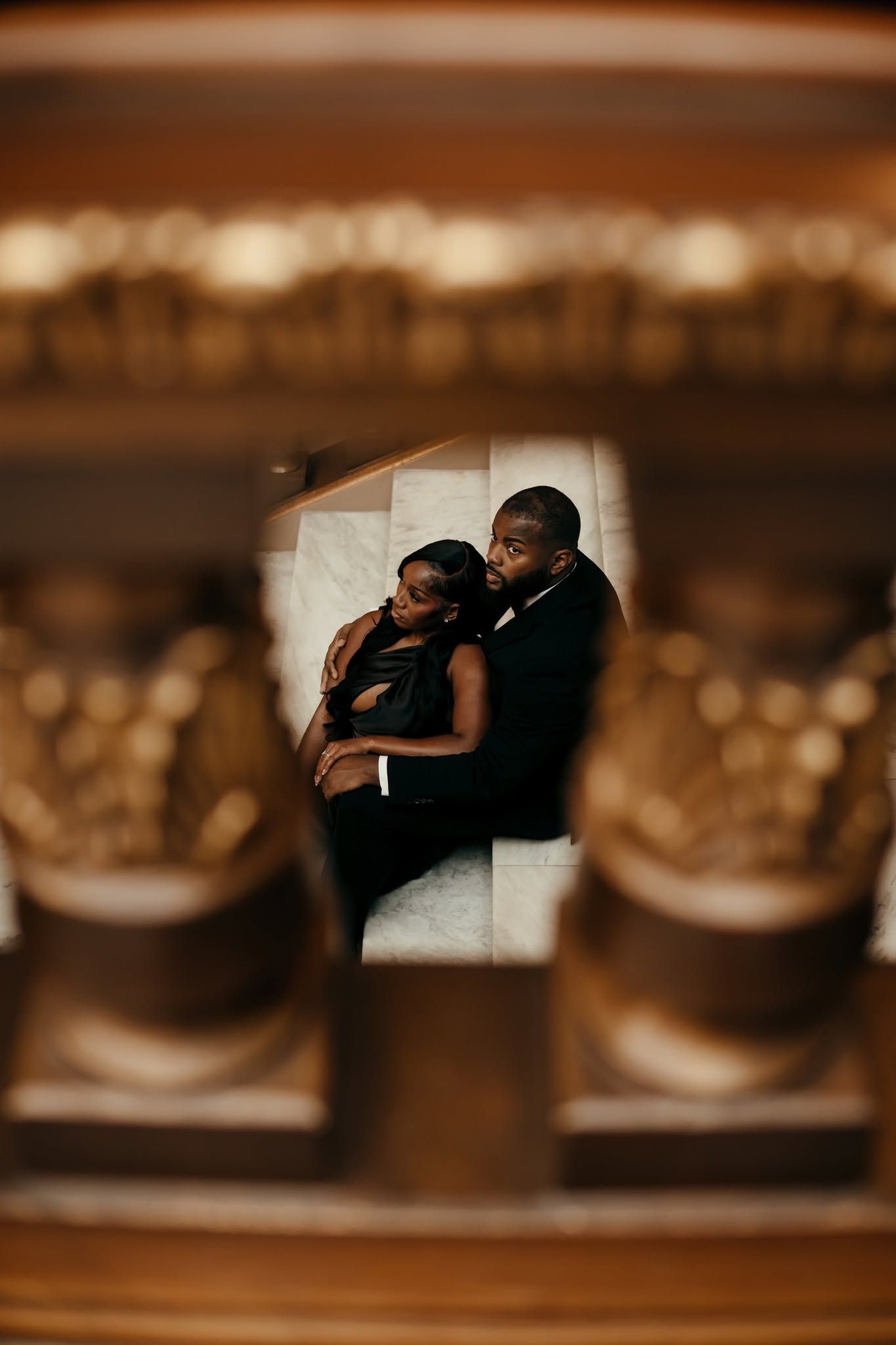 Elegant Black couple in formal attire sharing an intimate moment, framed by ornate golden columns in a grand venue.