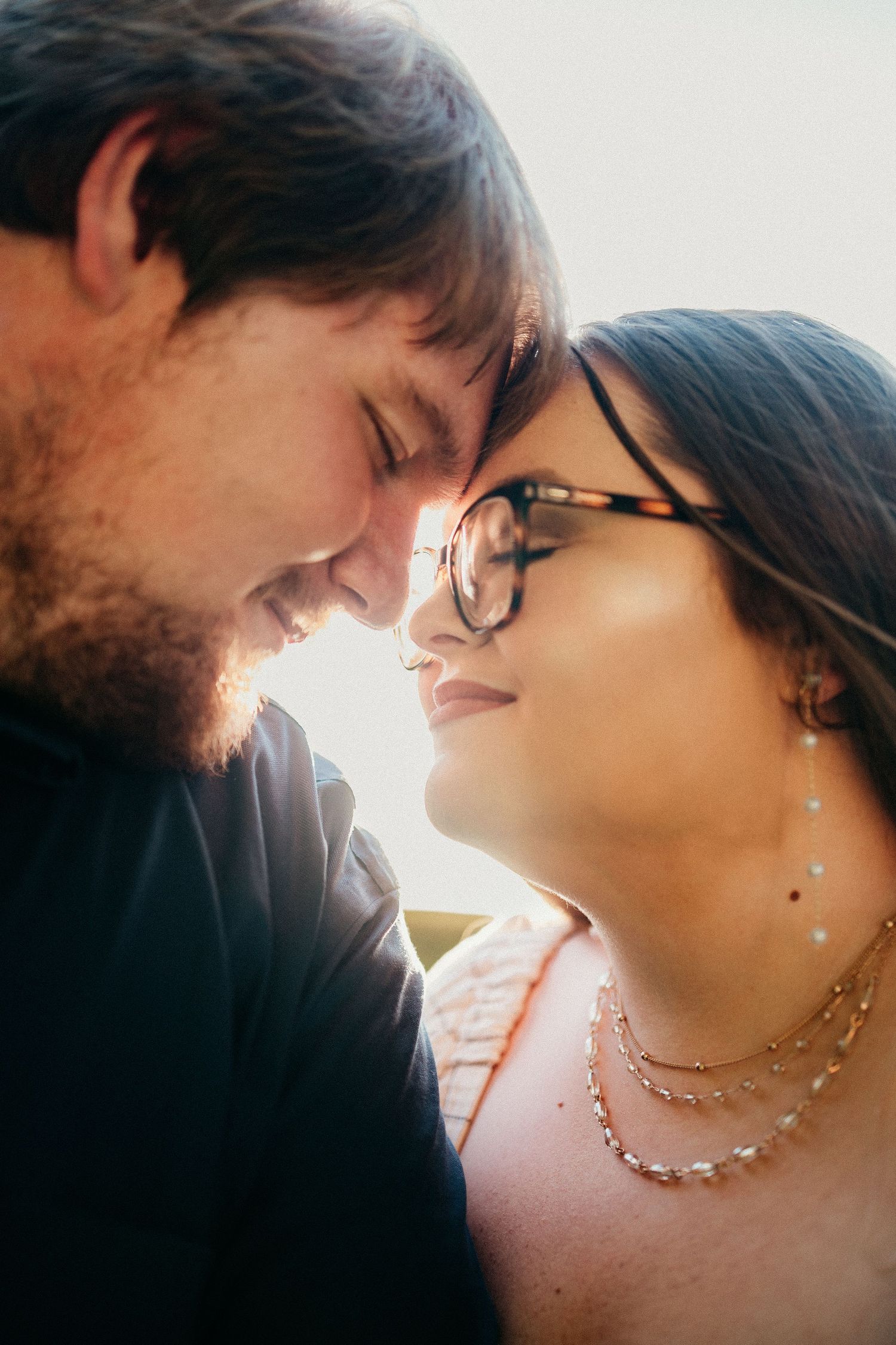 A smiling couple touching foreheads in a romantic close-up portrait with warm backlit glow, woman wearing glasses and layered necklaces.