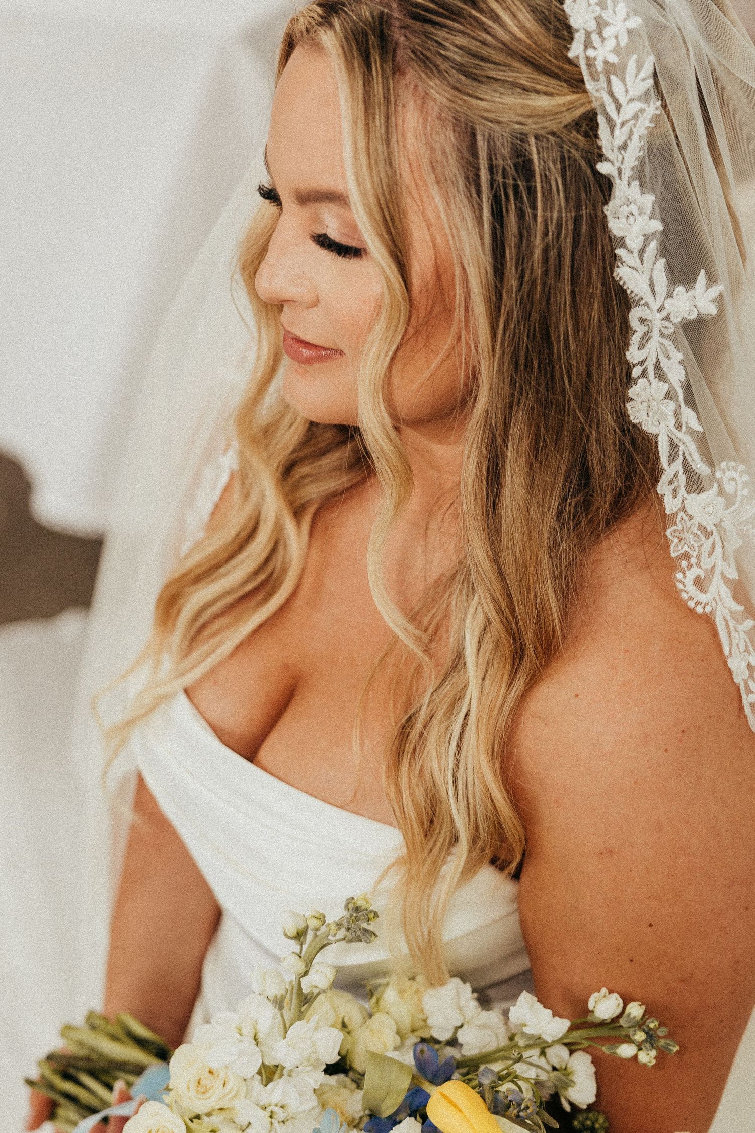 Blonde bride with lace-trimmed veil holds white and blue floral bouquet in profile view.