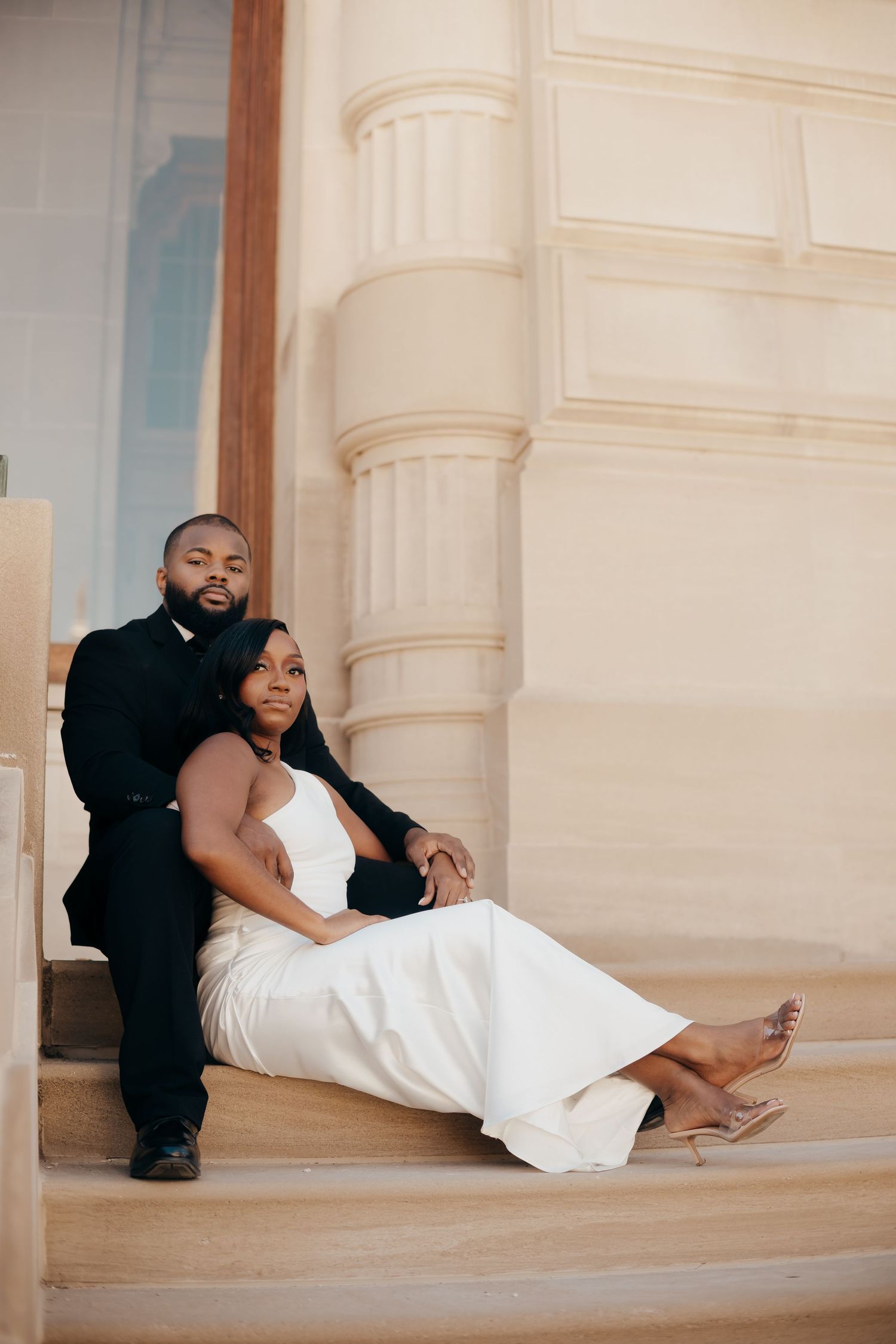 Couple in formal attire sitting close together on stone steps outside an elegant building during an engagement photo shoot.
