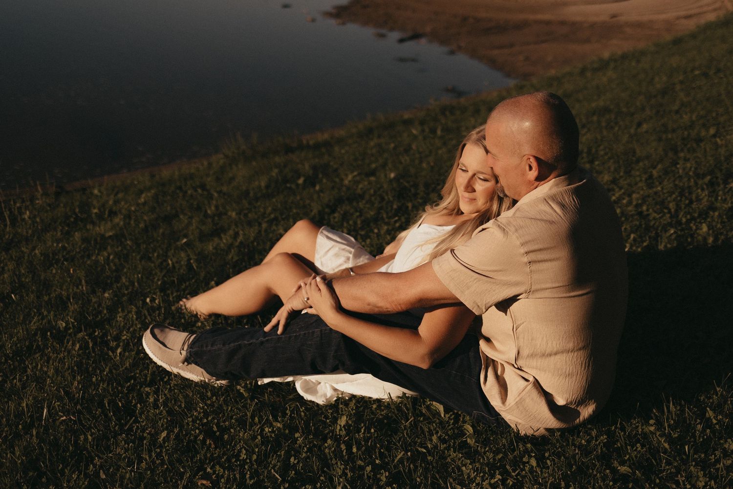 Couple sitting together on grass by a lake at sunset, sharing a romantic moment outdoors.