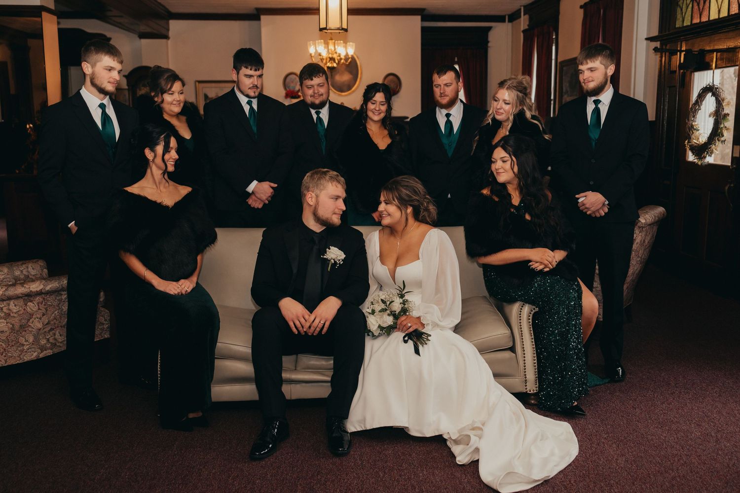 Wedding party in black attire surrounds bride and groom seated on elegant sofa in formal venue.