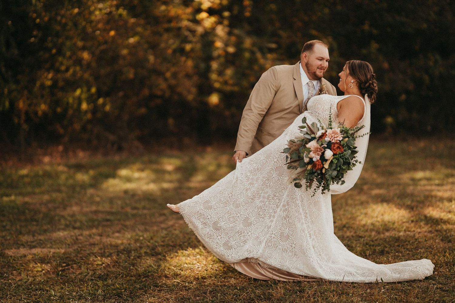 Groom in tan suit twirling bride in lace wedding gown while holding floral bouquet outdoors.