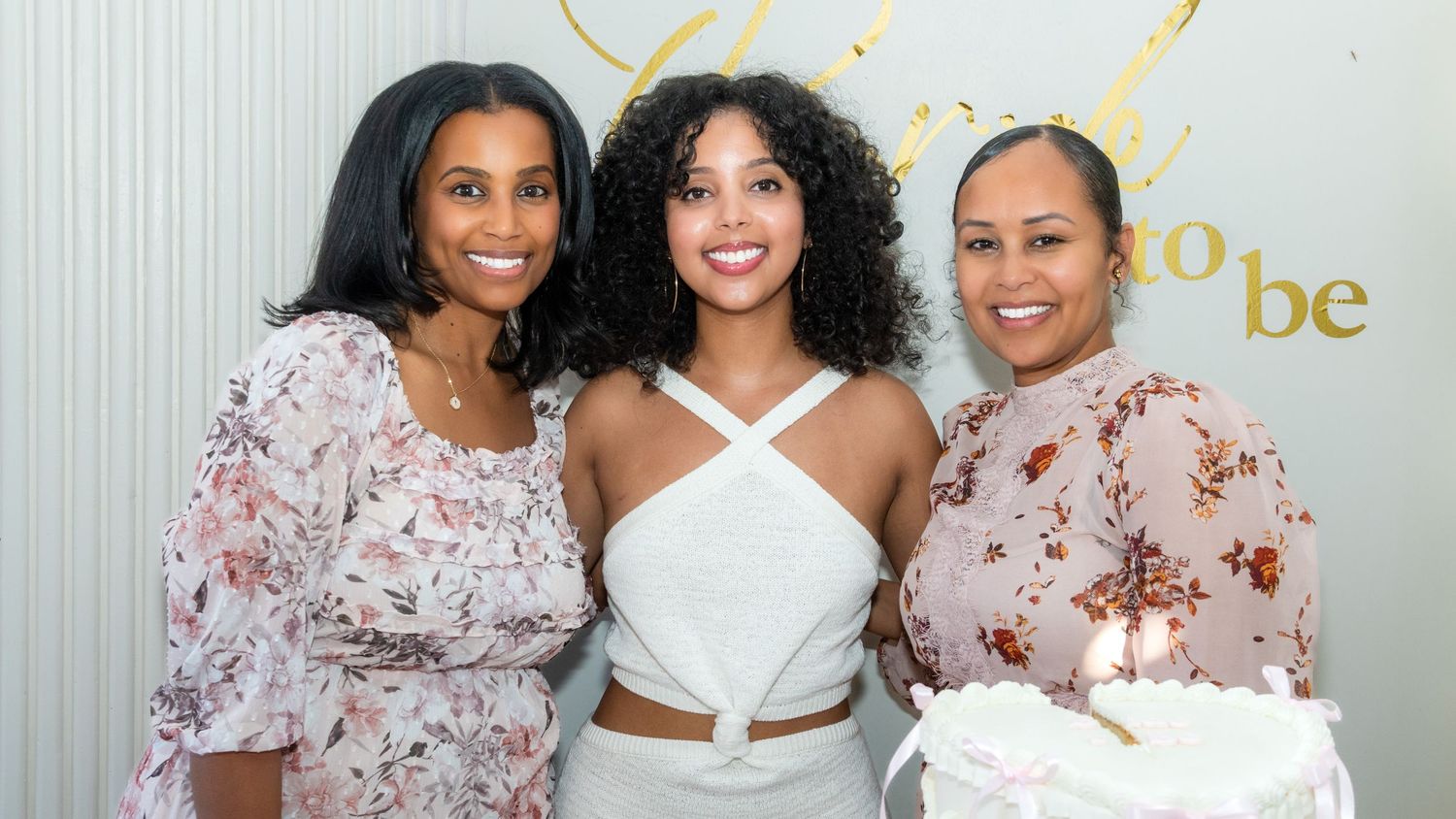Three women posing together at a bridal shower celebration with a cake