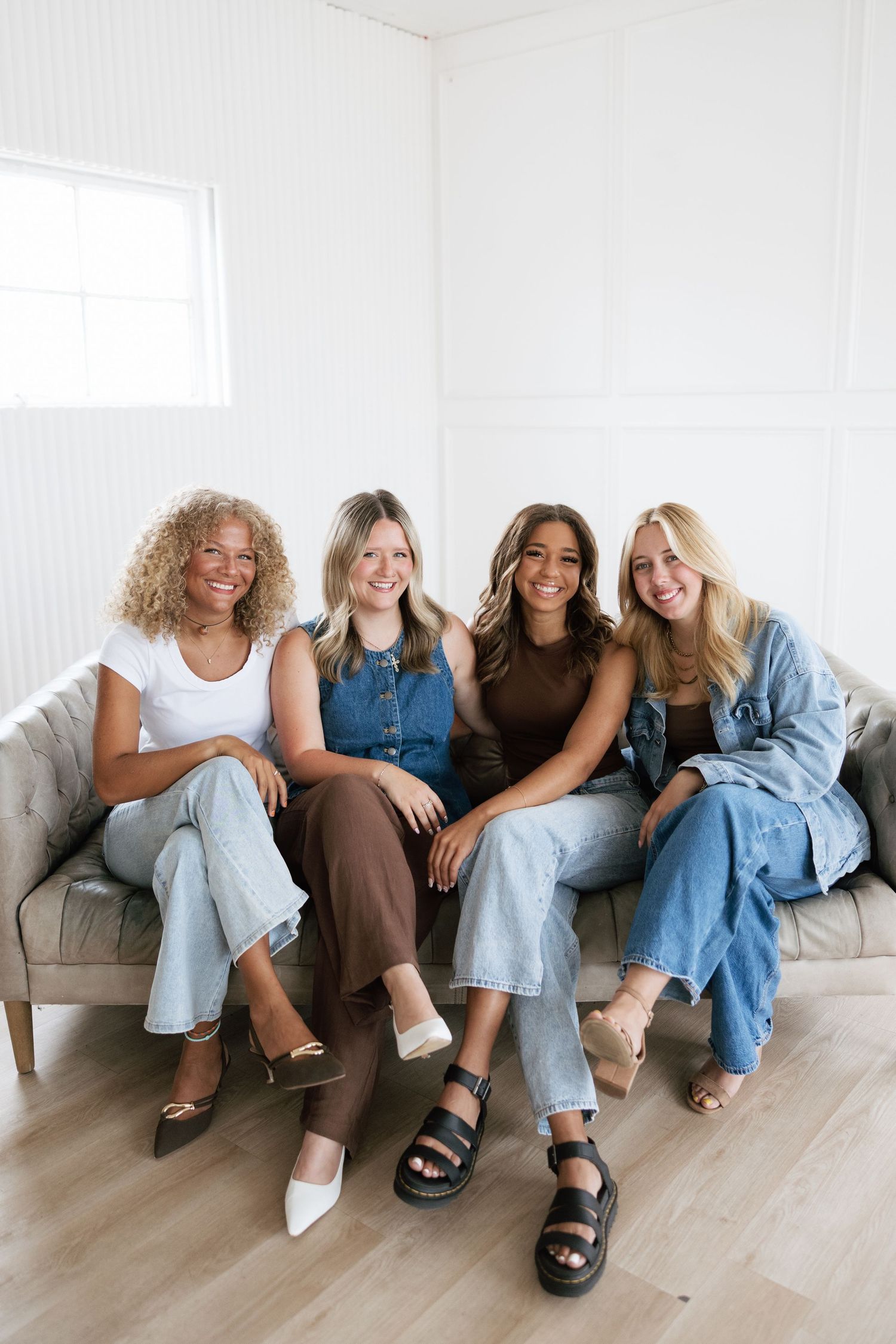 Four smiling women sitting together on a gray couch in a bright white room, wearing casual denim and neutral outfits.