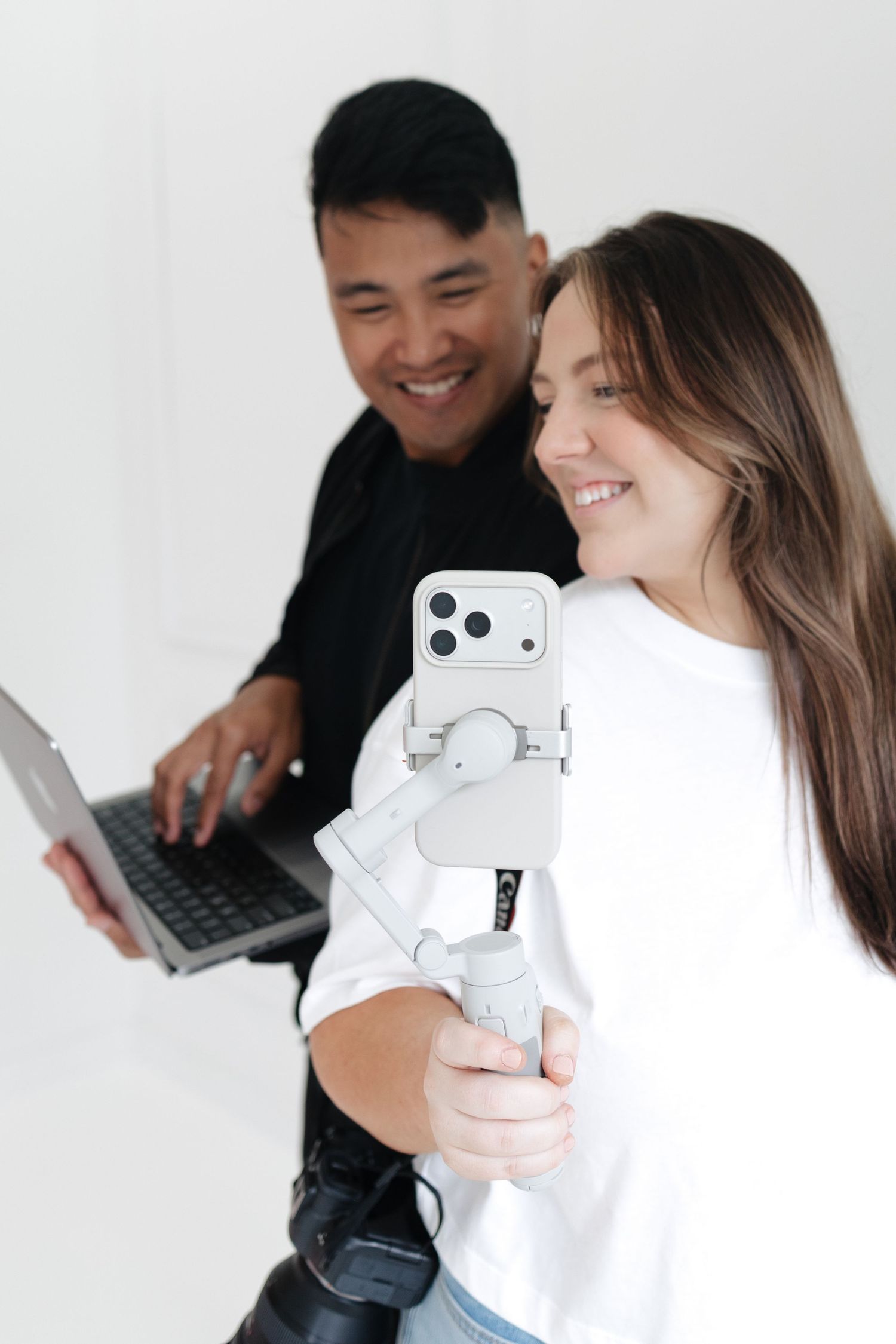 A smiling woman holds a smartphone on a gimbal stabilizer while a man with a laptop poses behind her on white background.