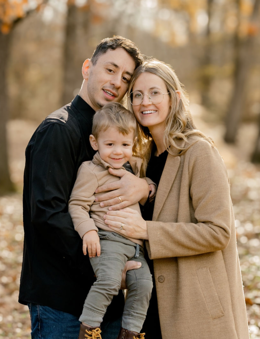 Happy family with young toddler boy posing together in autumn forest with golden fall foliage.