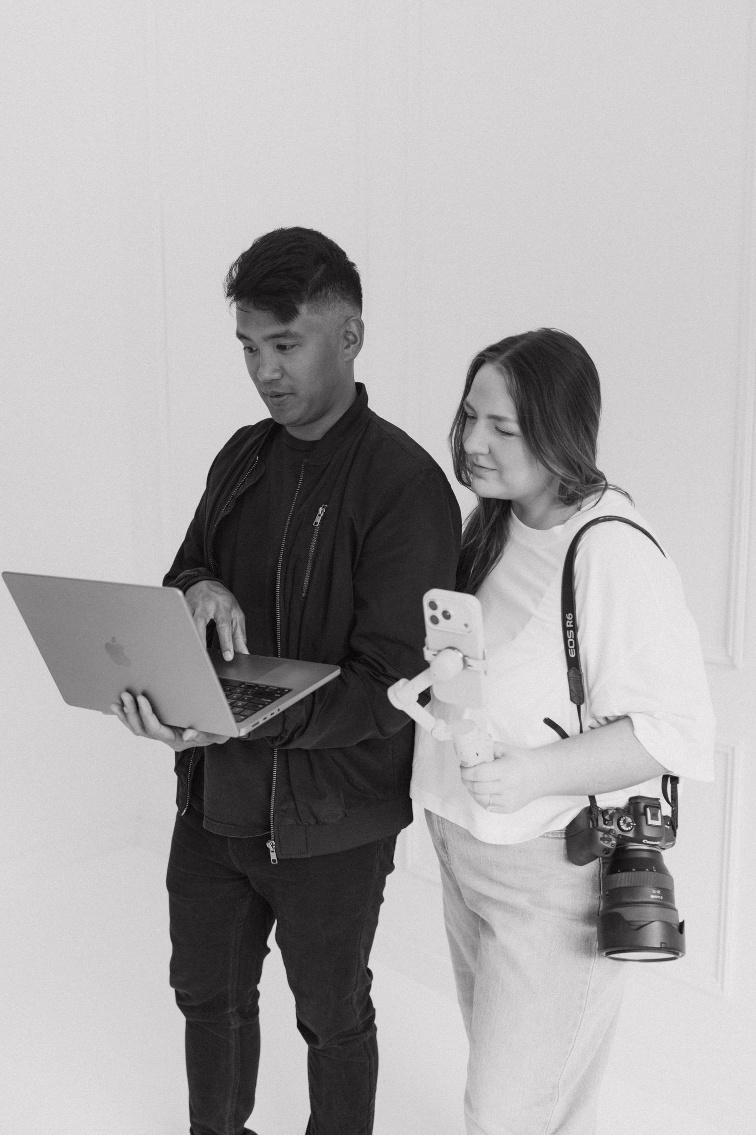 Two photographers review images on a laptop in a studio, woman holding DSLR camera and smartphone.