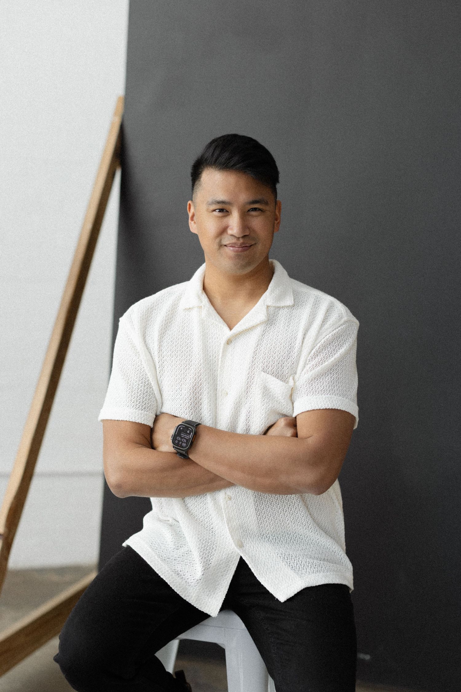 Smiling Asian man in white polo shirt sitting with arms crossed against gray and white background.