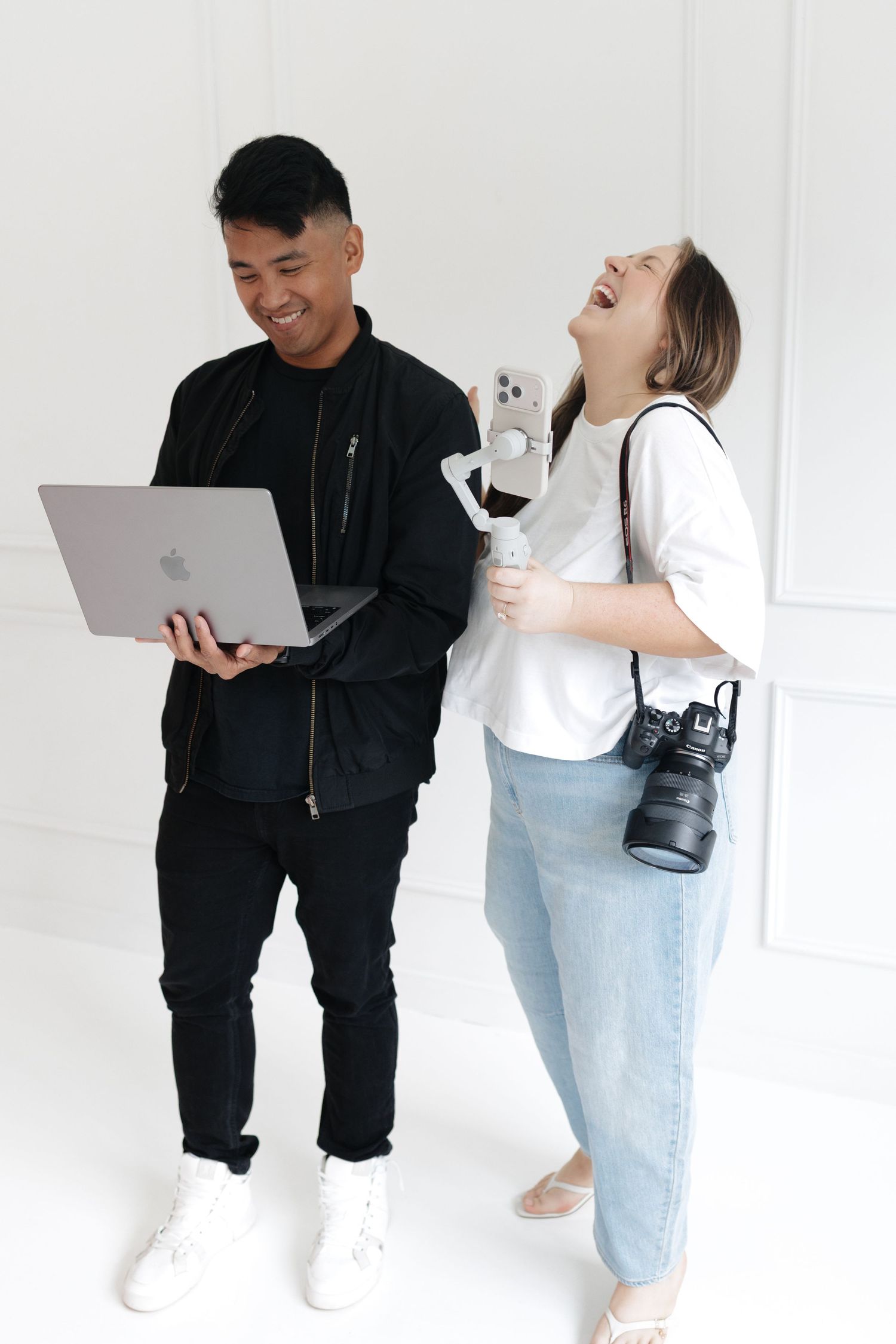 Two content creators laughing together, one holding a MacBook laptop and the other holding a camera and smartphone gimbal.