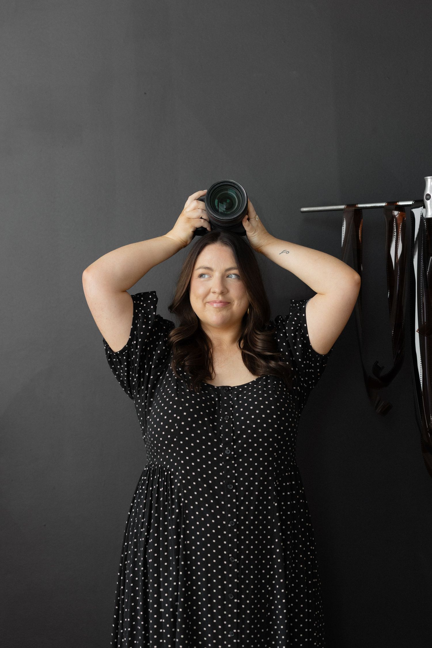 Plus size woman in polka dot dress holding camera above head in dark studio with backdrop stand.