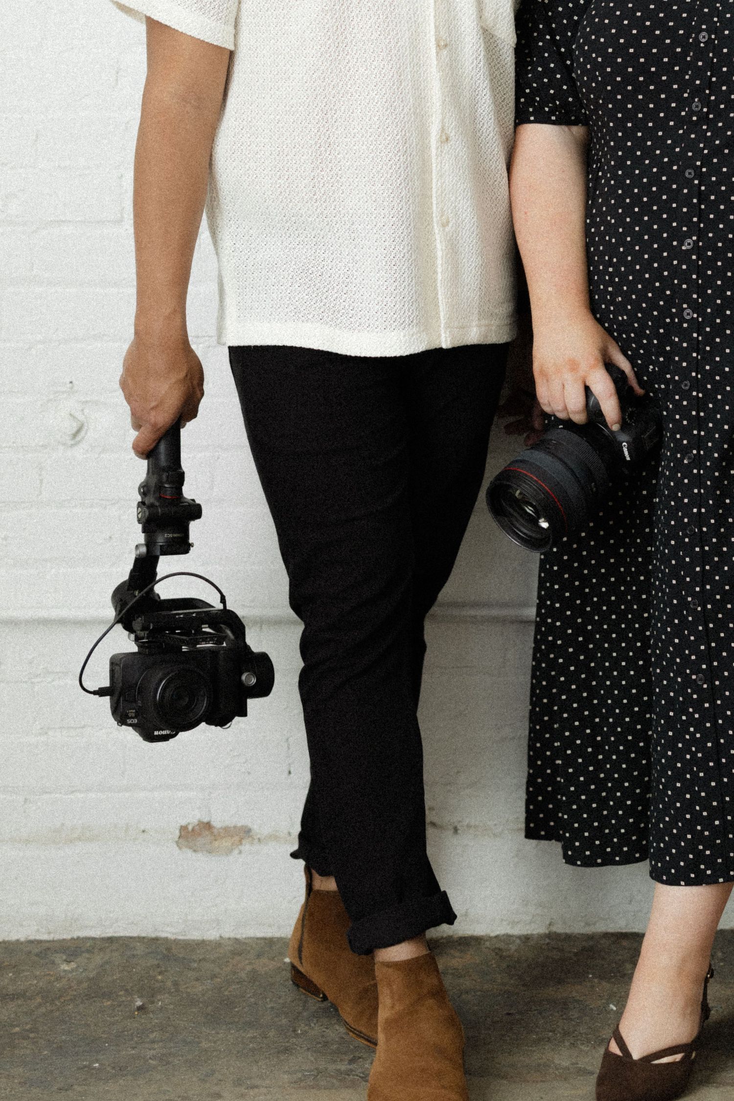 Two photographers standing side by side holding camera gimbal and DSLR camera against white brick wall.