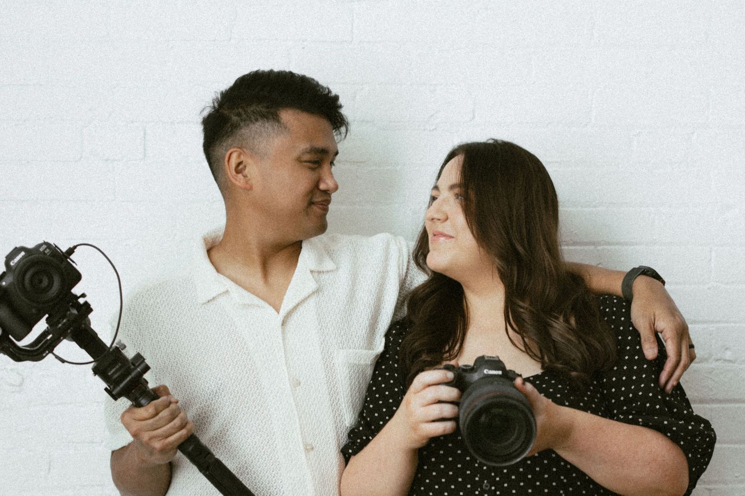 Man and woman photographers smiling at each other holding camera and gimbal against white brick wall.