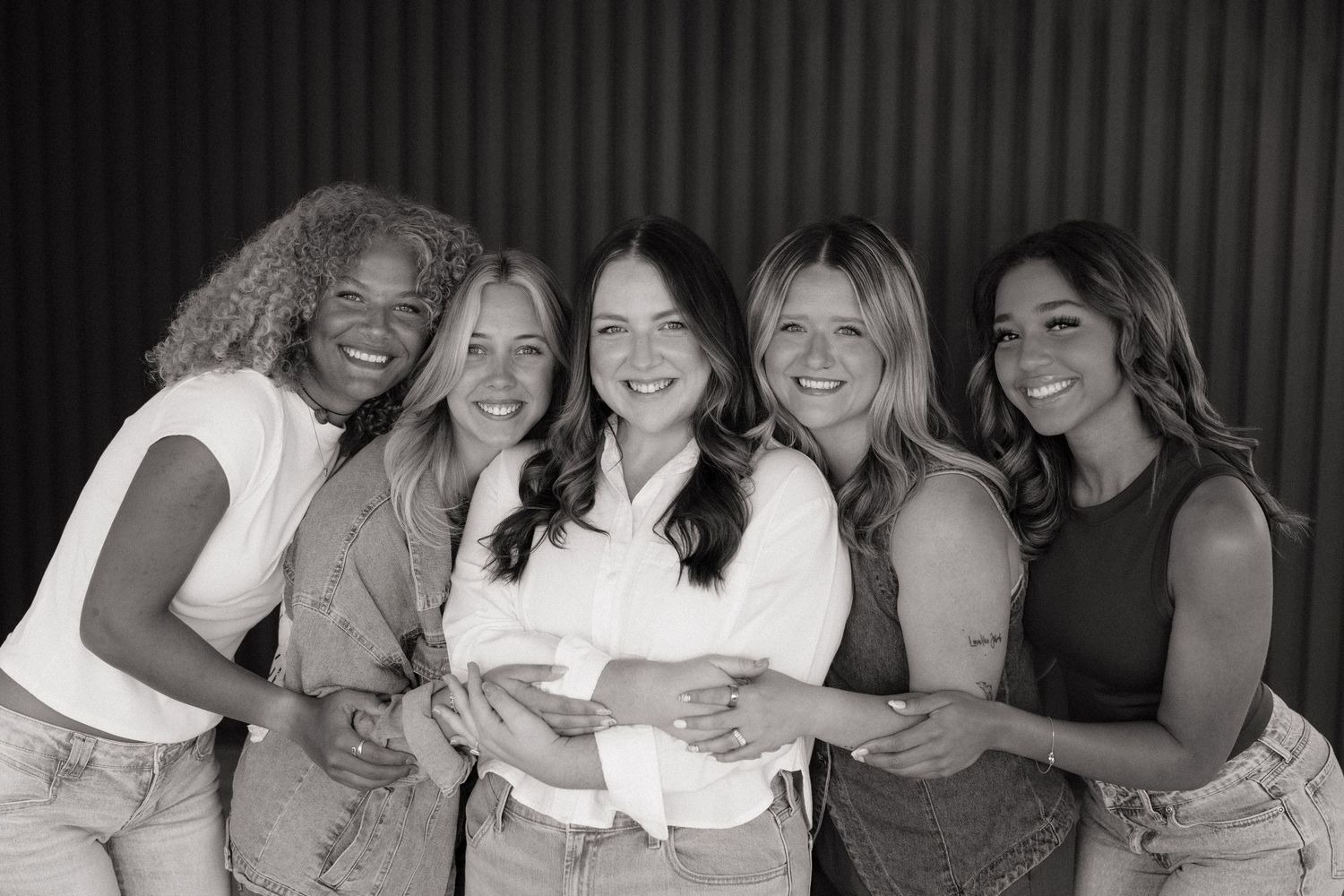 Five women smiling and posing together in a black and white group portrait photo.