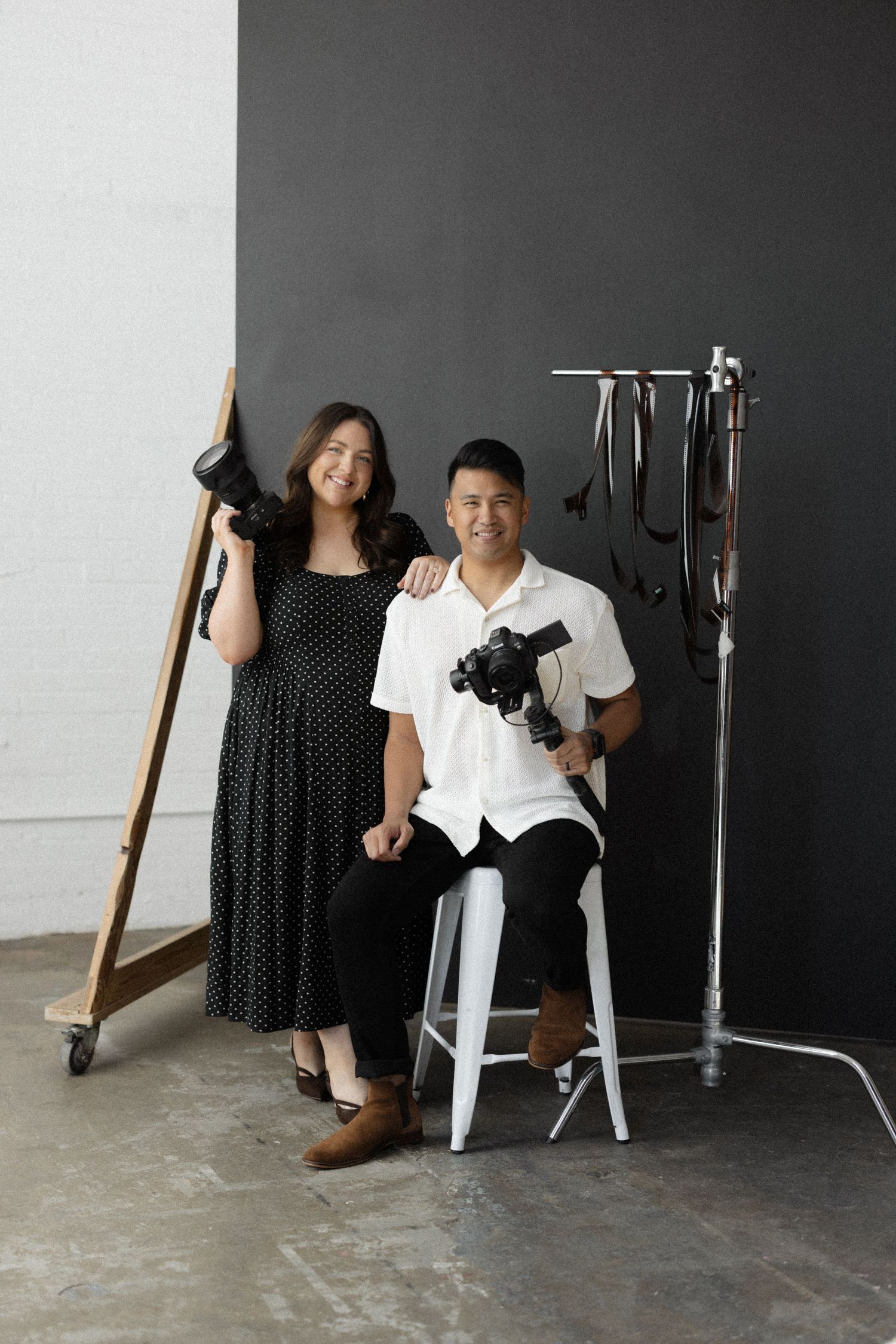 A smiling couple poses in a photography studio, holding cameras and equipment against a dark gray backdrop.