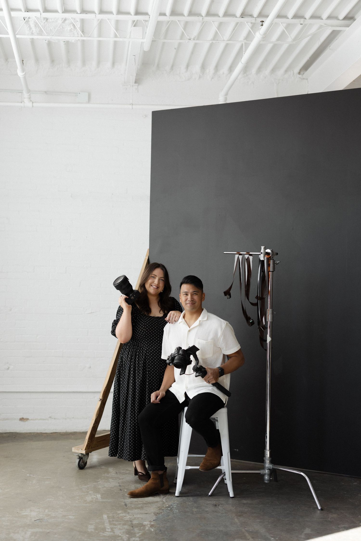 A man and woman holding cameras pose in a professional photography studio with dark backdrop.