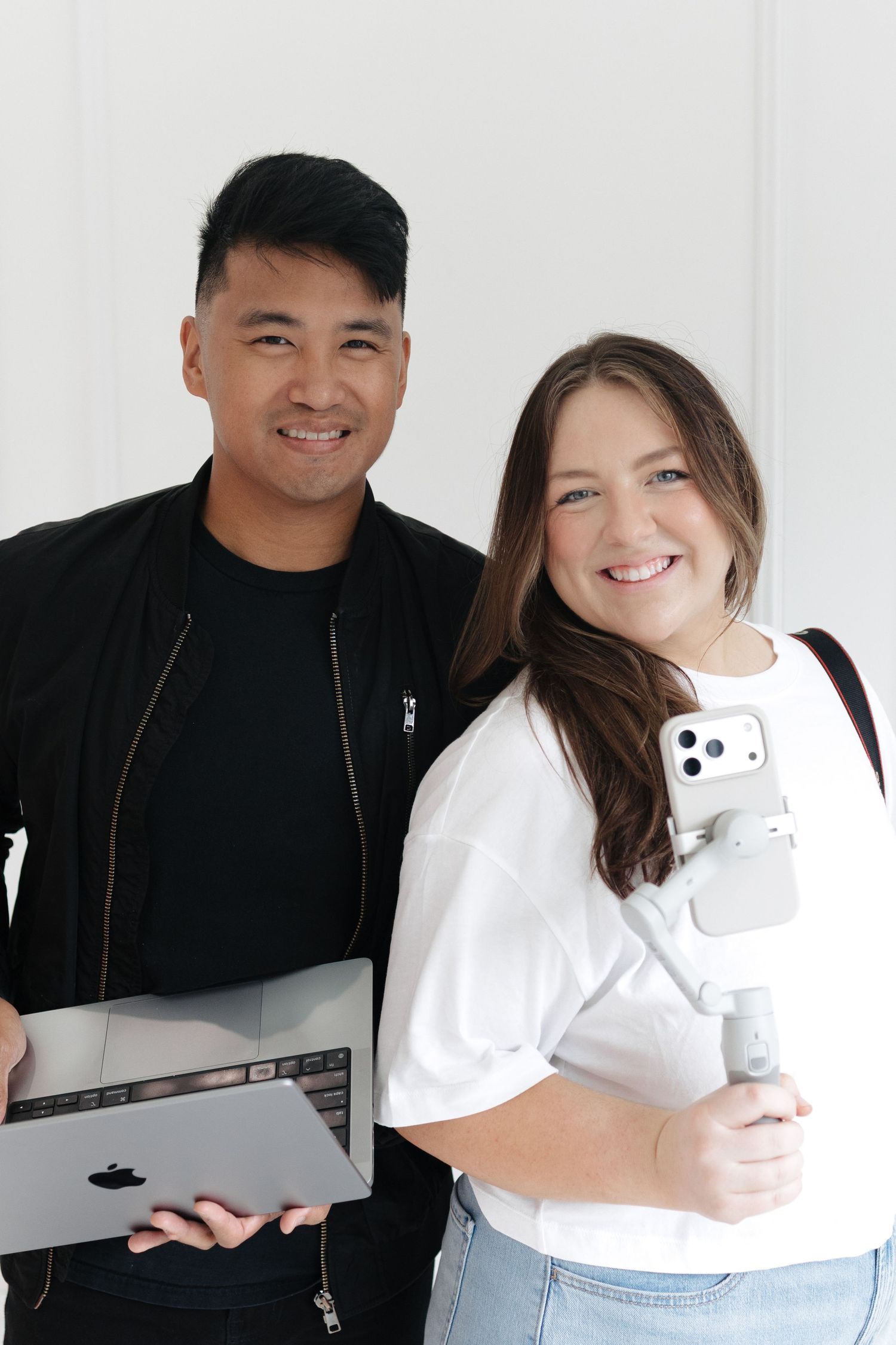 Two smiling content creators pose together, one holding a MacBook and the other a smartphone gimbal stabilizer.