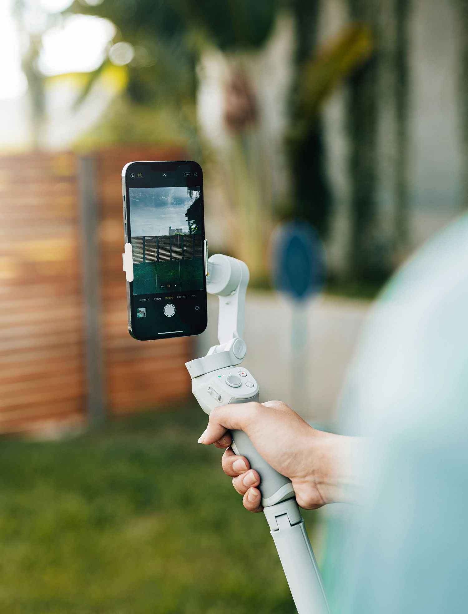 Person holding smartphone on DJI OM gimbal stabilizer outdoors in garden setting.