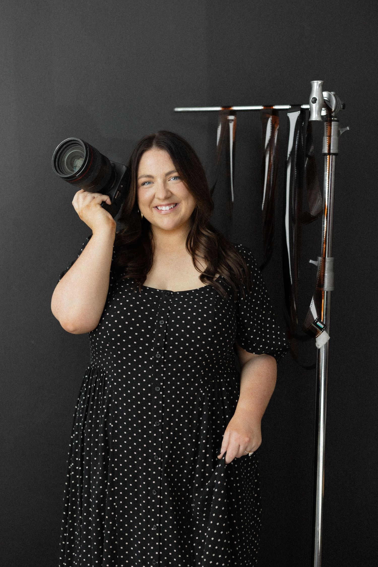 Smiling woman in polka dot dress holding camera on shoulder beside backdrop stand in dark studio.